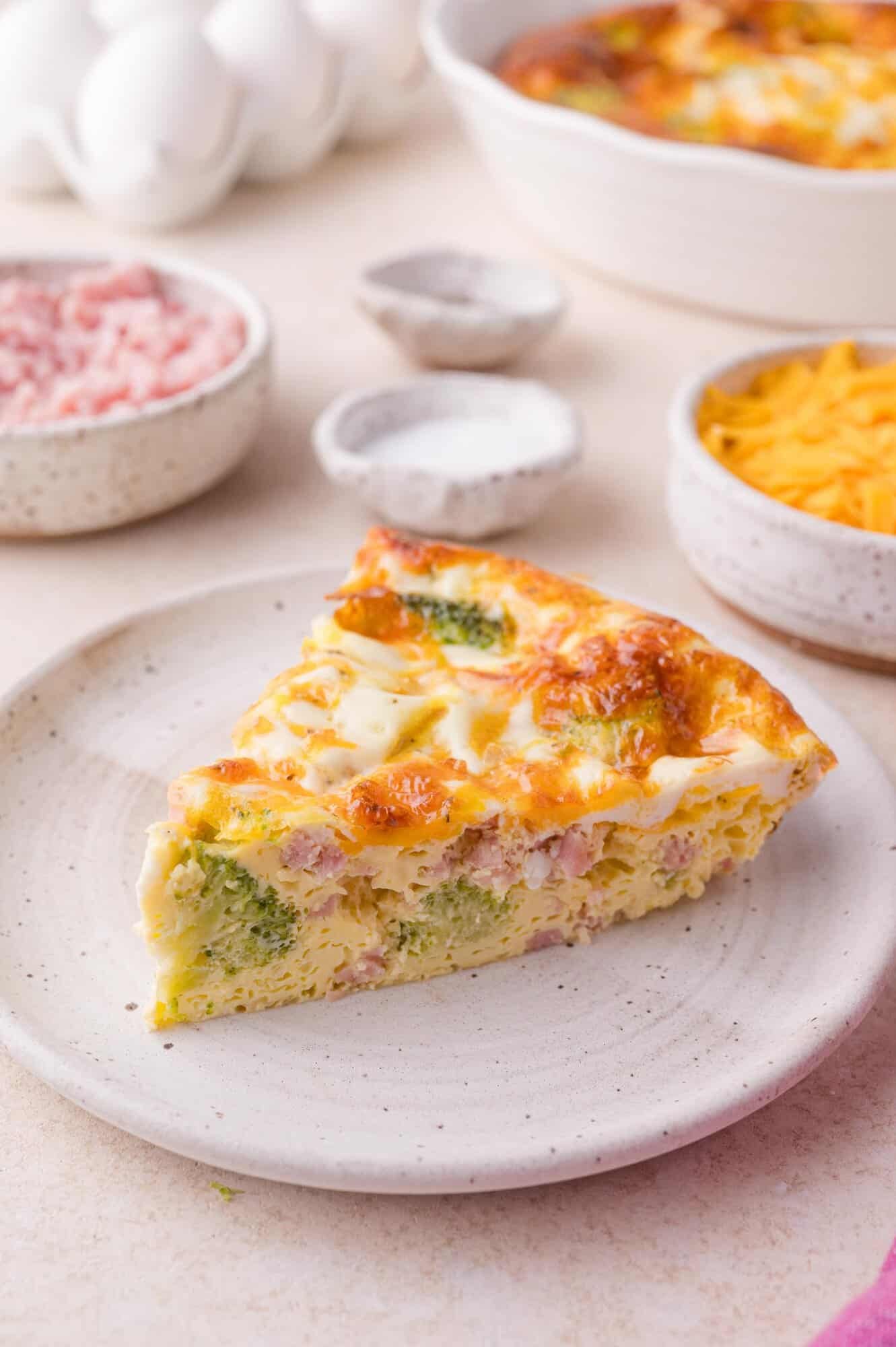 A slice of broccoli, ham, and cheese quiche sits on a white plate, with bowls of diced ham, shredded cheese, and eggs in the background.