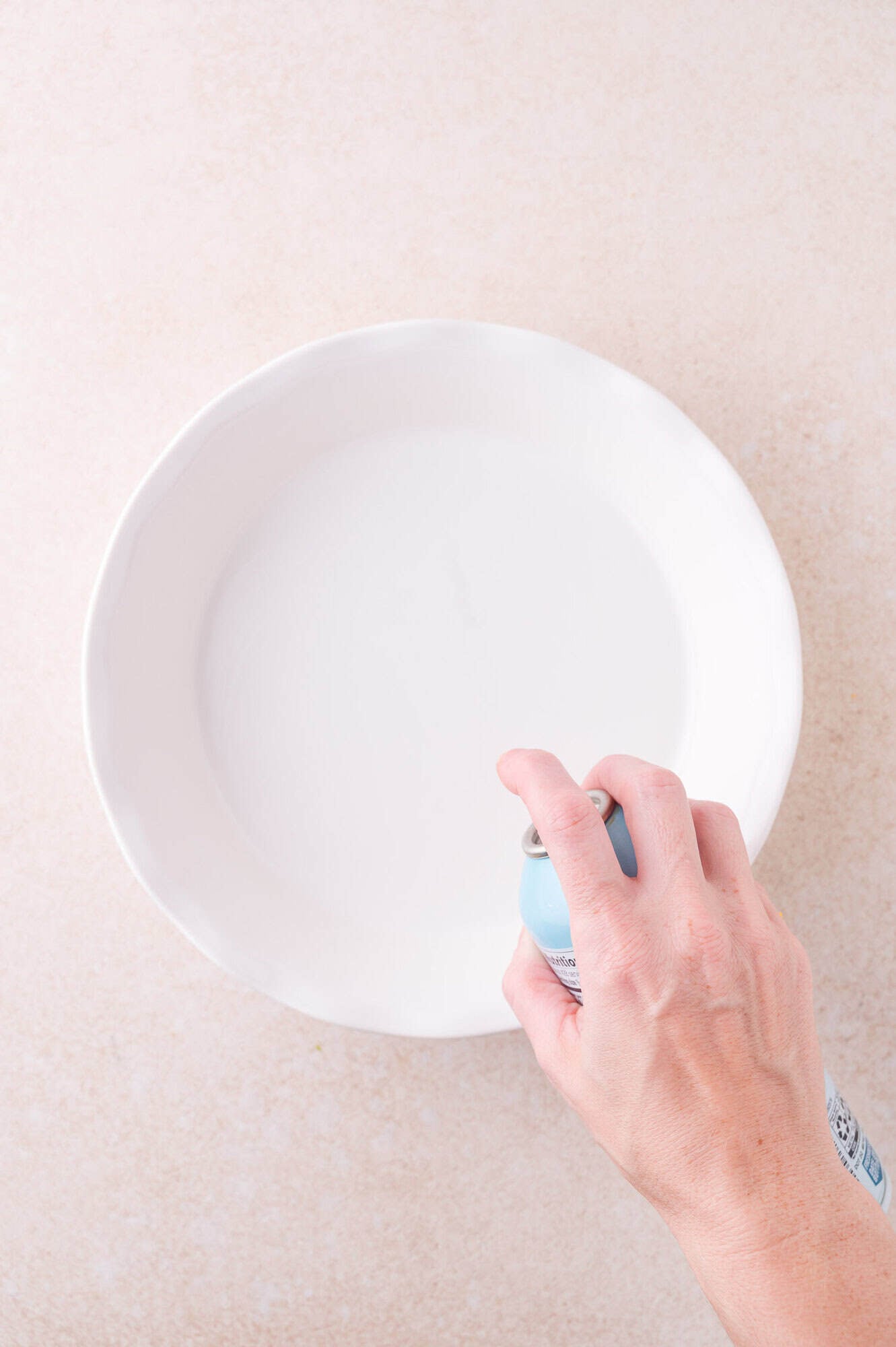 A hand sprays nonstick cooking spray into a round, white ceramic pie dish on a beige surface.