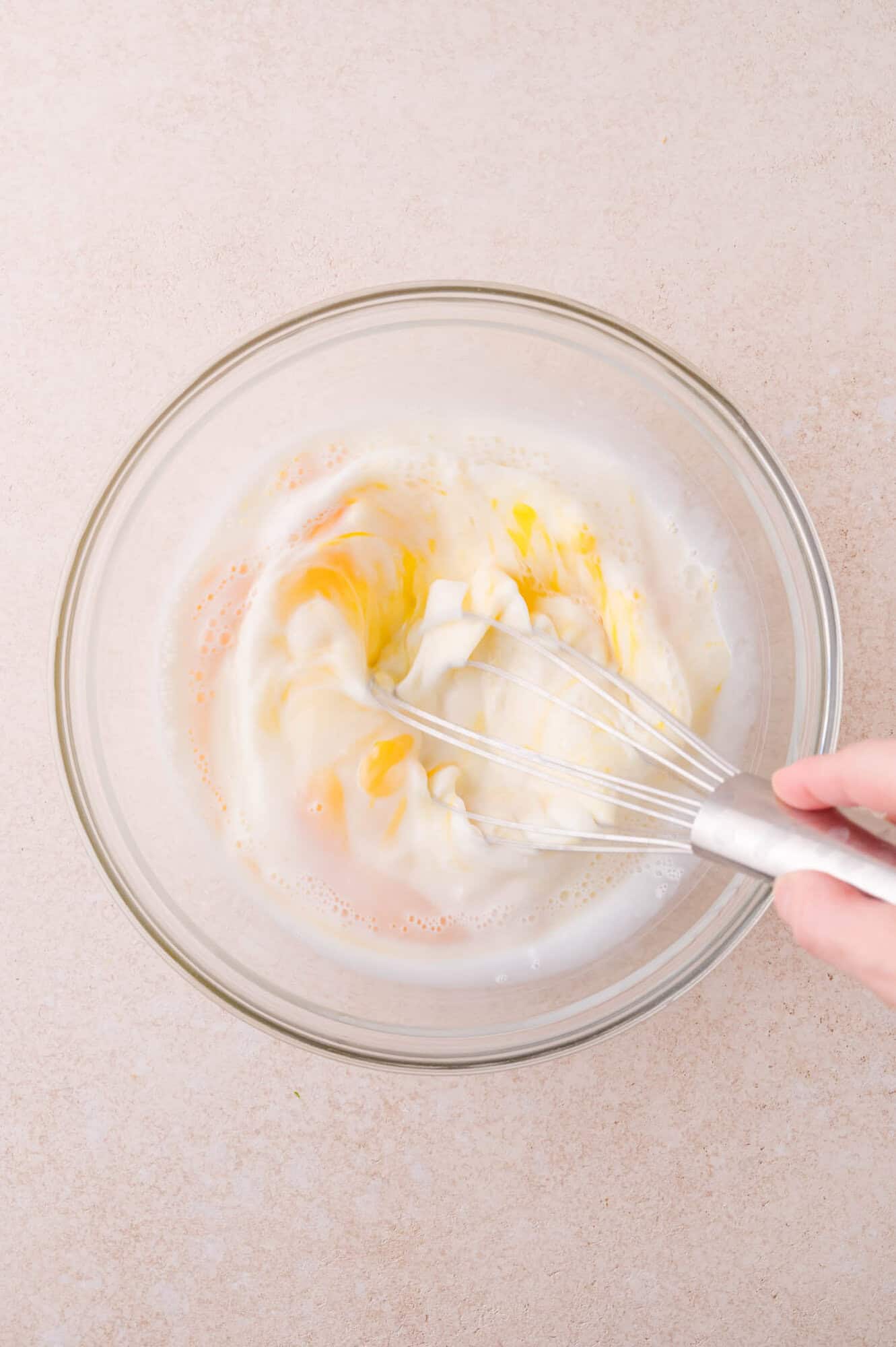 A hand uses a metal whisk to mix eggs and milk in a clear glass bowl on a light-colored countertop.