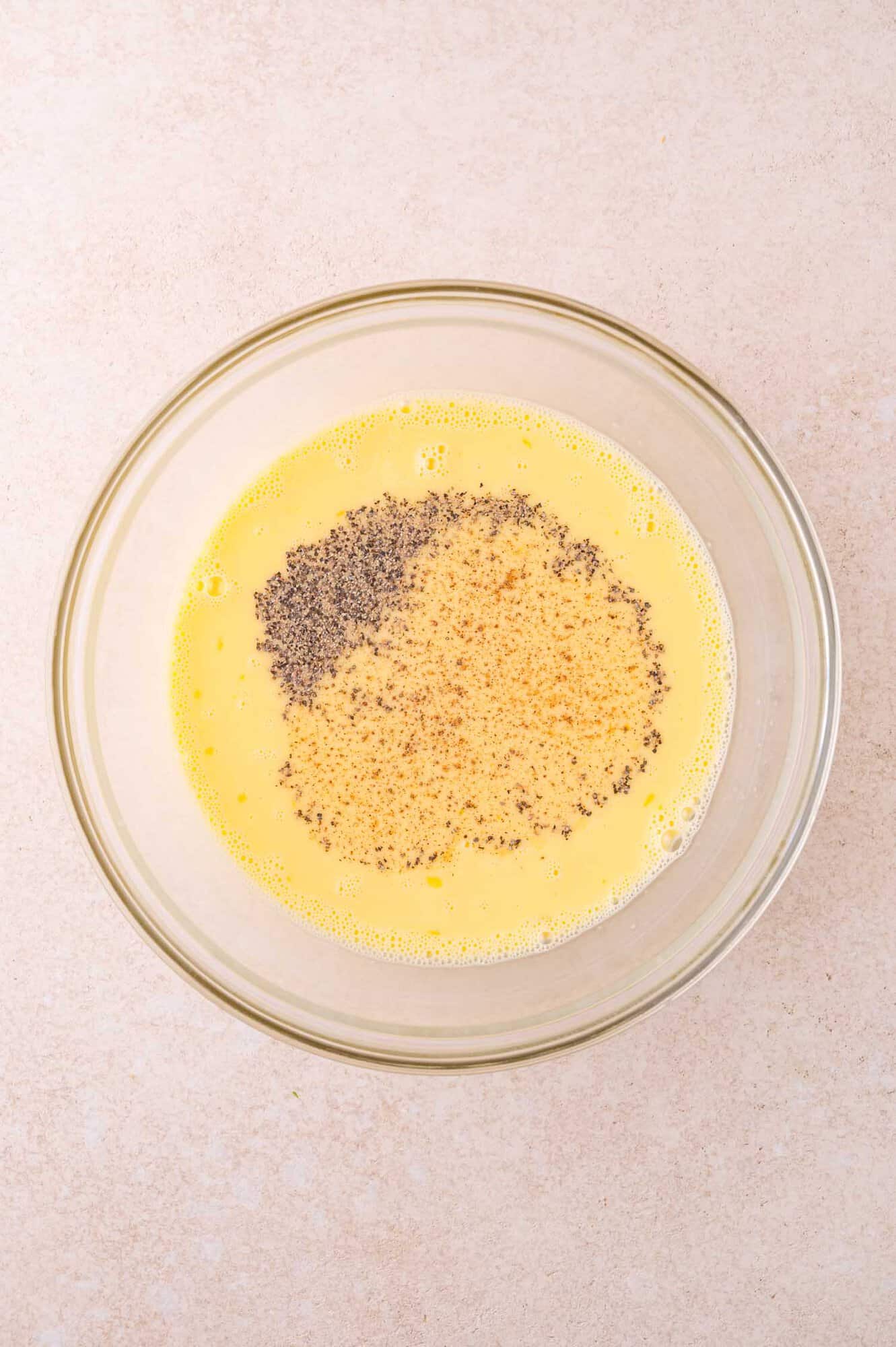 A clear glass bowl contains beaten eggs with black and tan seasonings on top, photographed from above on a light-colored surface.