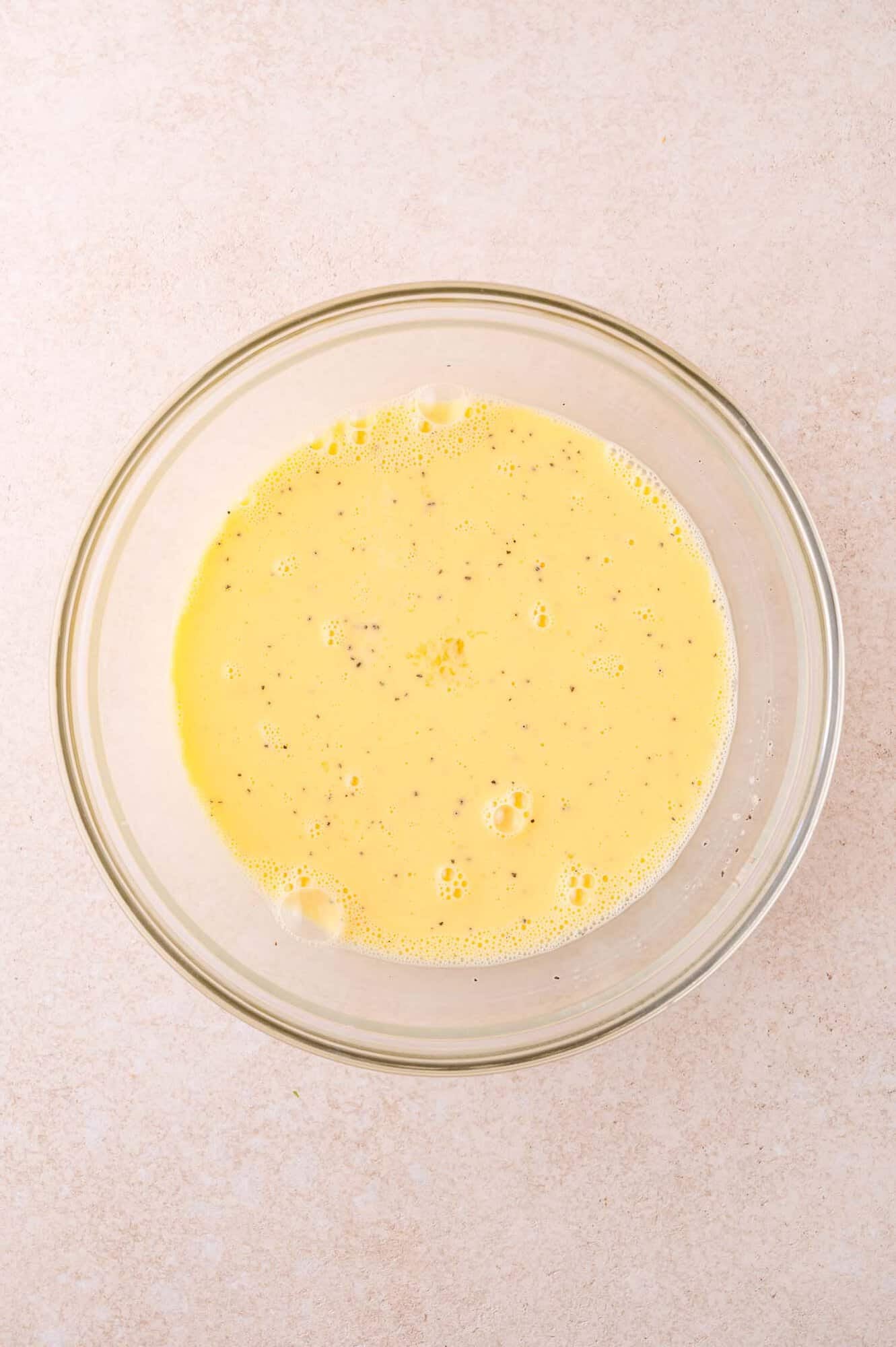 A glass bowl filled with a whisked egg mixture, speckled with black pepper, sits on a light-colored countertop.