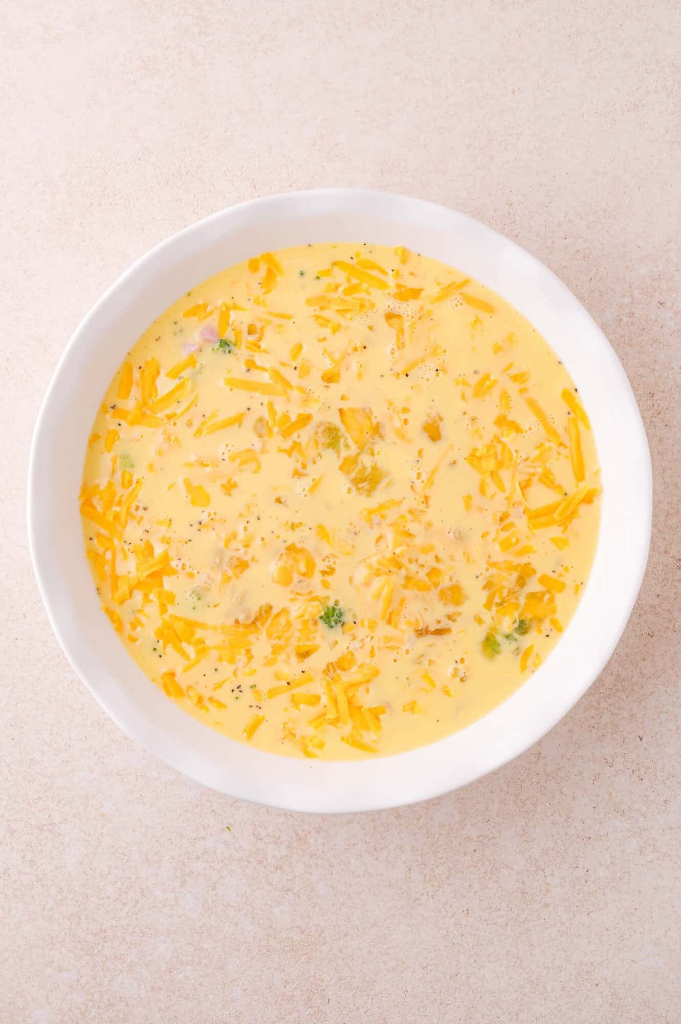 A white bowl filled with a yellow egg mixture containing shredded cheese and small green vegetable pieces, on a light surface.