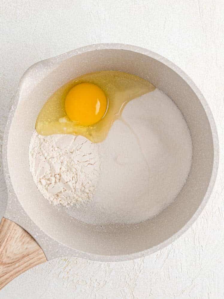 A saucepan with flour, sugar, and a raw egg on a light surface.