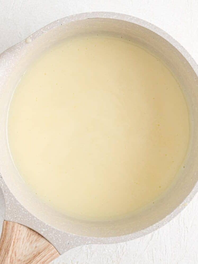 A saucepan filled with a smooth, pale yellow vanilla pudding sits on a light textured surface.