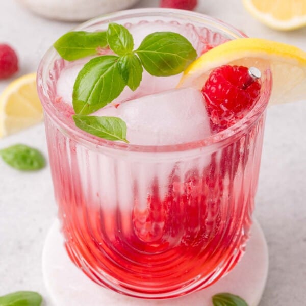 Glass of raspberry beverage with ice, a lemon slice, raspberry, and fresh basil on top, placed on a white coaster with lemon slices and basil leaves in the background.