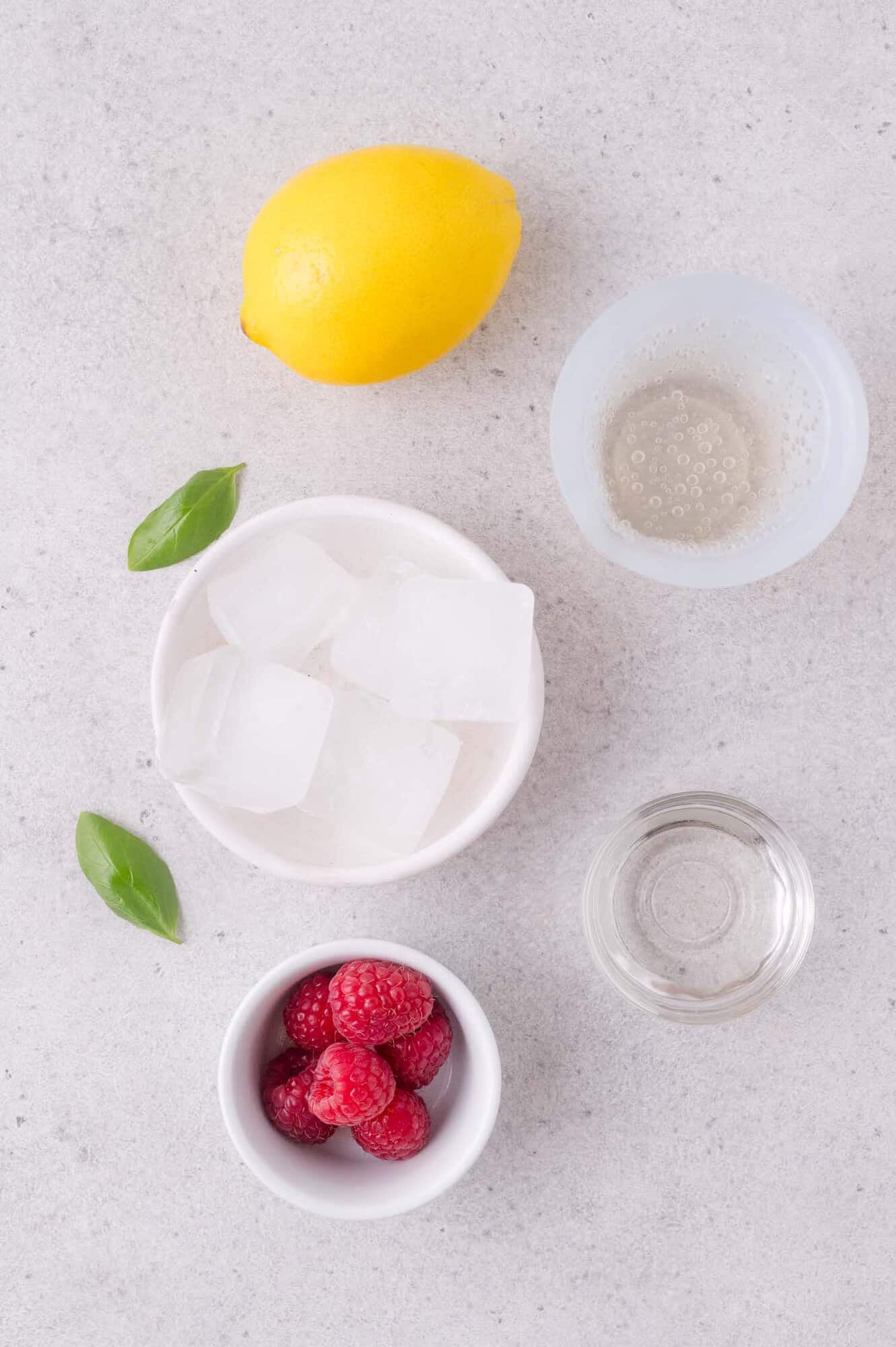 Lemon, glass of club soda, basil leaves, bowl of ice cubes, bowl of raspberries, and a small glass of clear liquid on a light surface.