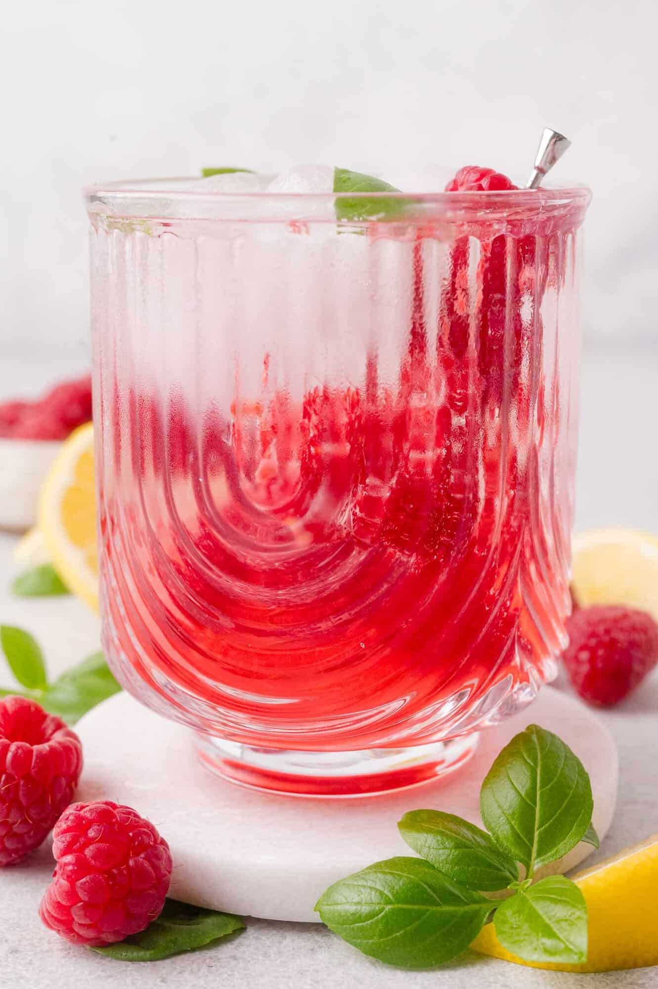A glass of iced raspberry mocktail garnished with fresh raspberries, basil leaves, and a metal straw, surrounded by lemon slices and basil on a white surface.