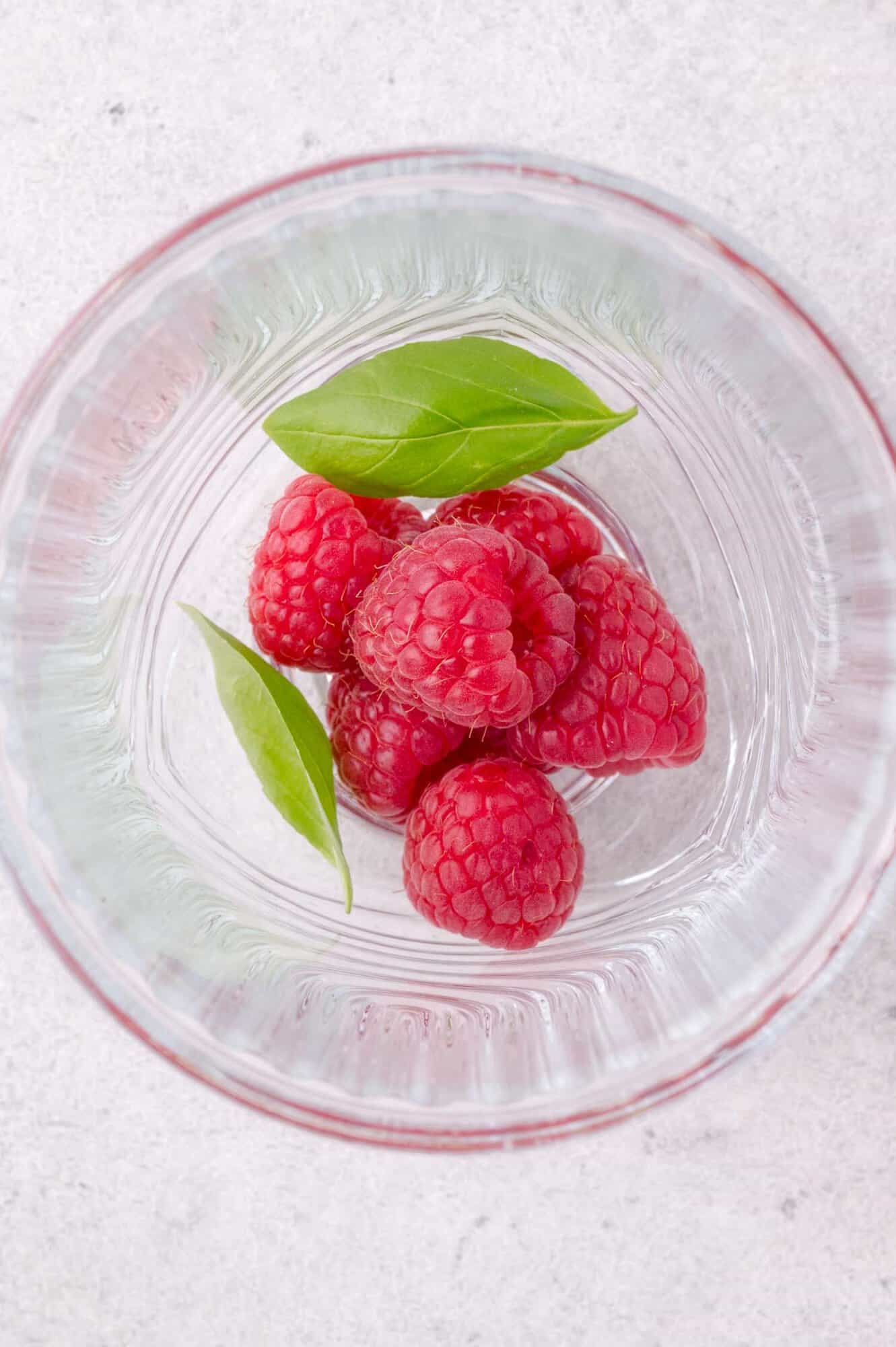 Five fresh raspberries and two green basil leaves in the bottom of a clear glass, viewed from above on a light surface.