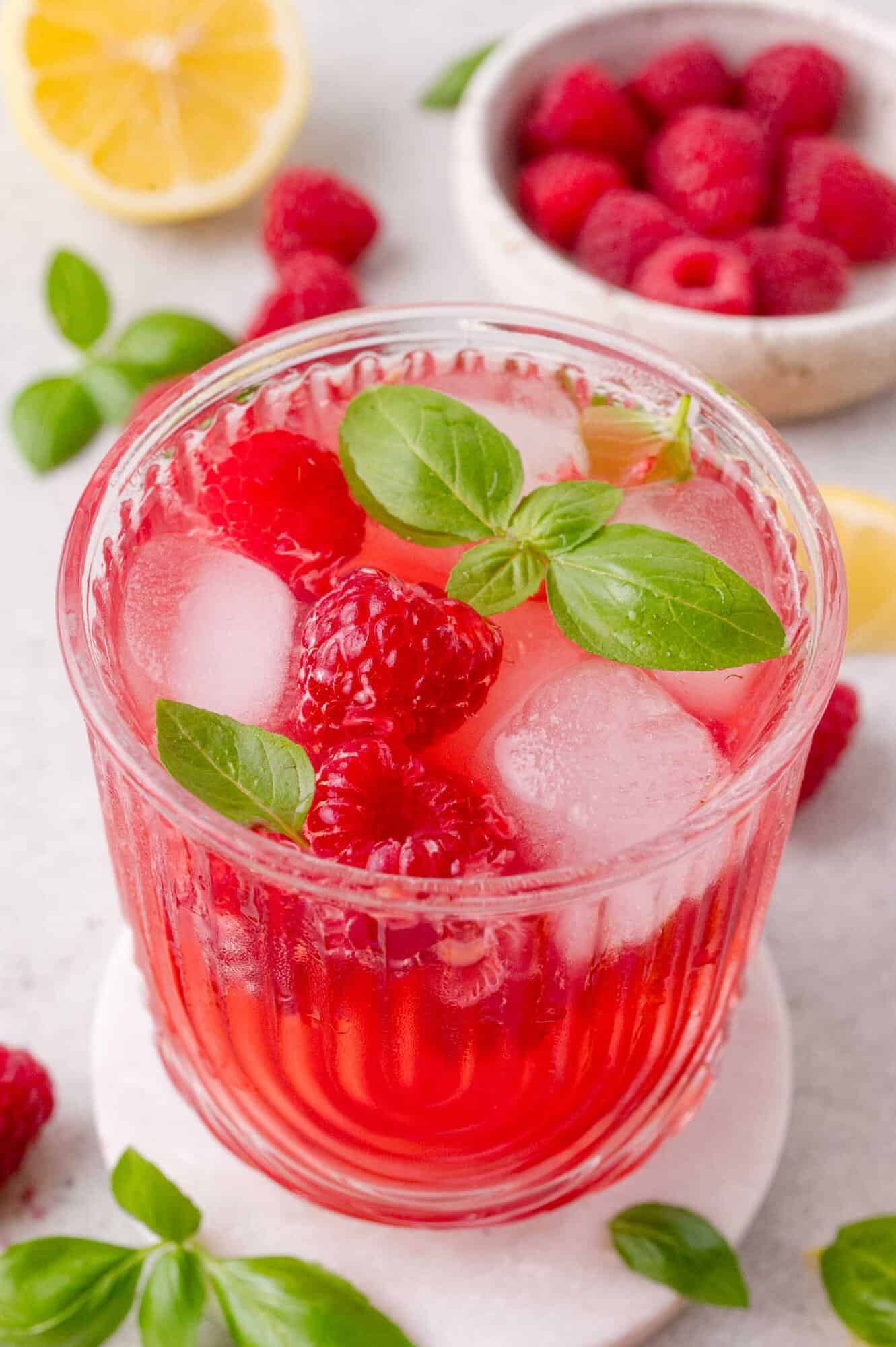 A glass of dark pink raspberry smash mocktail with ice cubes, fresh raspberries, and basil leaves, placed on a white coaster with raspberries and a lemon half in the background.