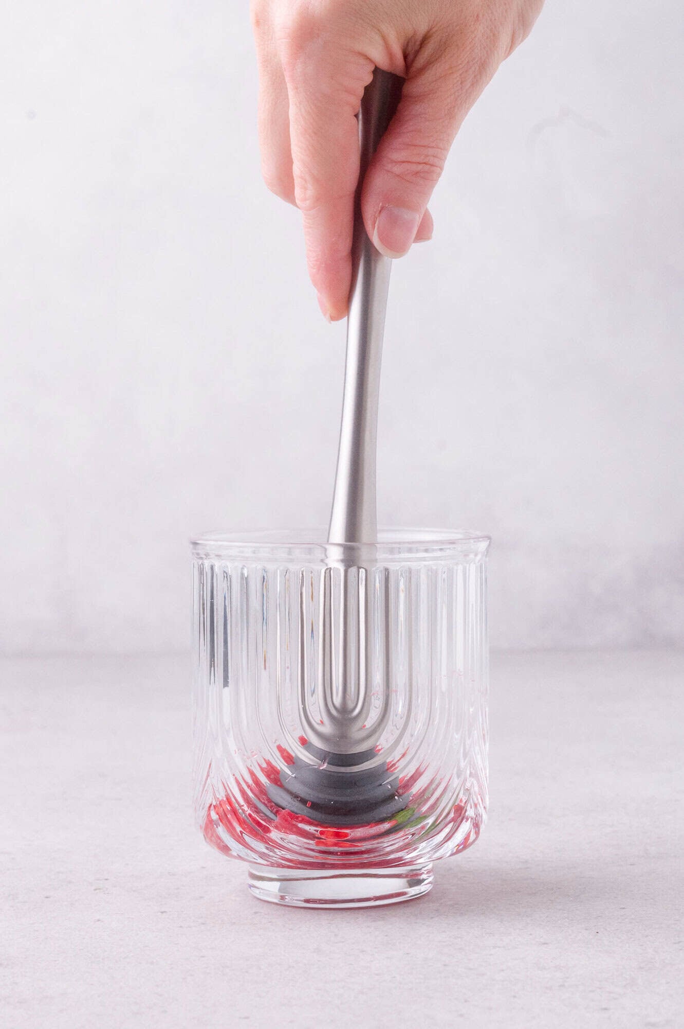 A hand uses a metal muddler to mash red berries at the bottom of a clear, textured glass on a gray surface.