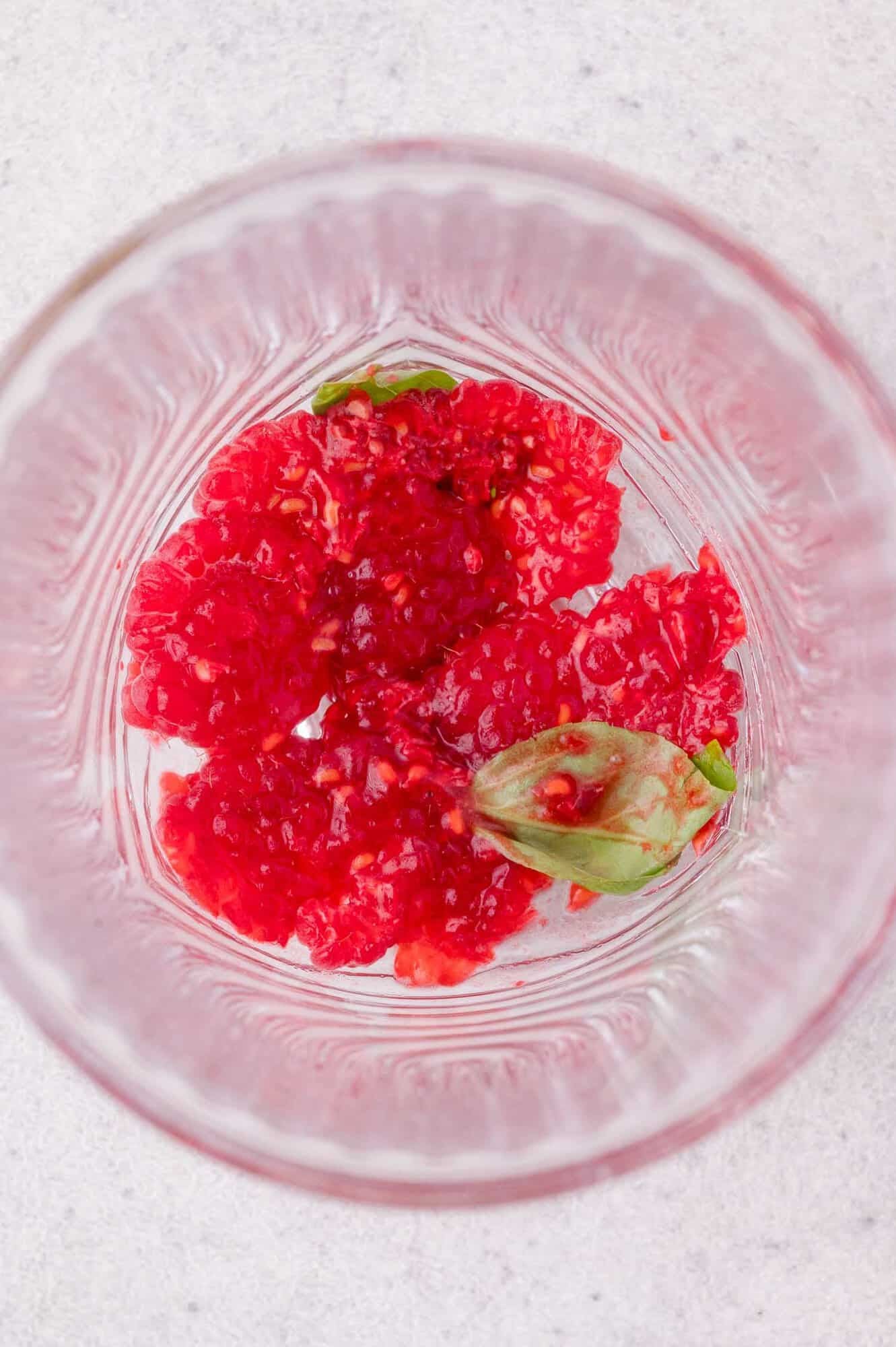 Crushed raspberries and a few basil leaves at the bottom of a clear glass on a light-colored surface.
