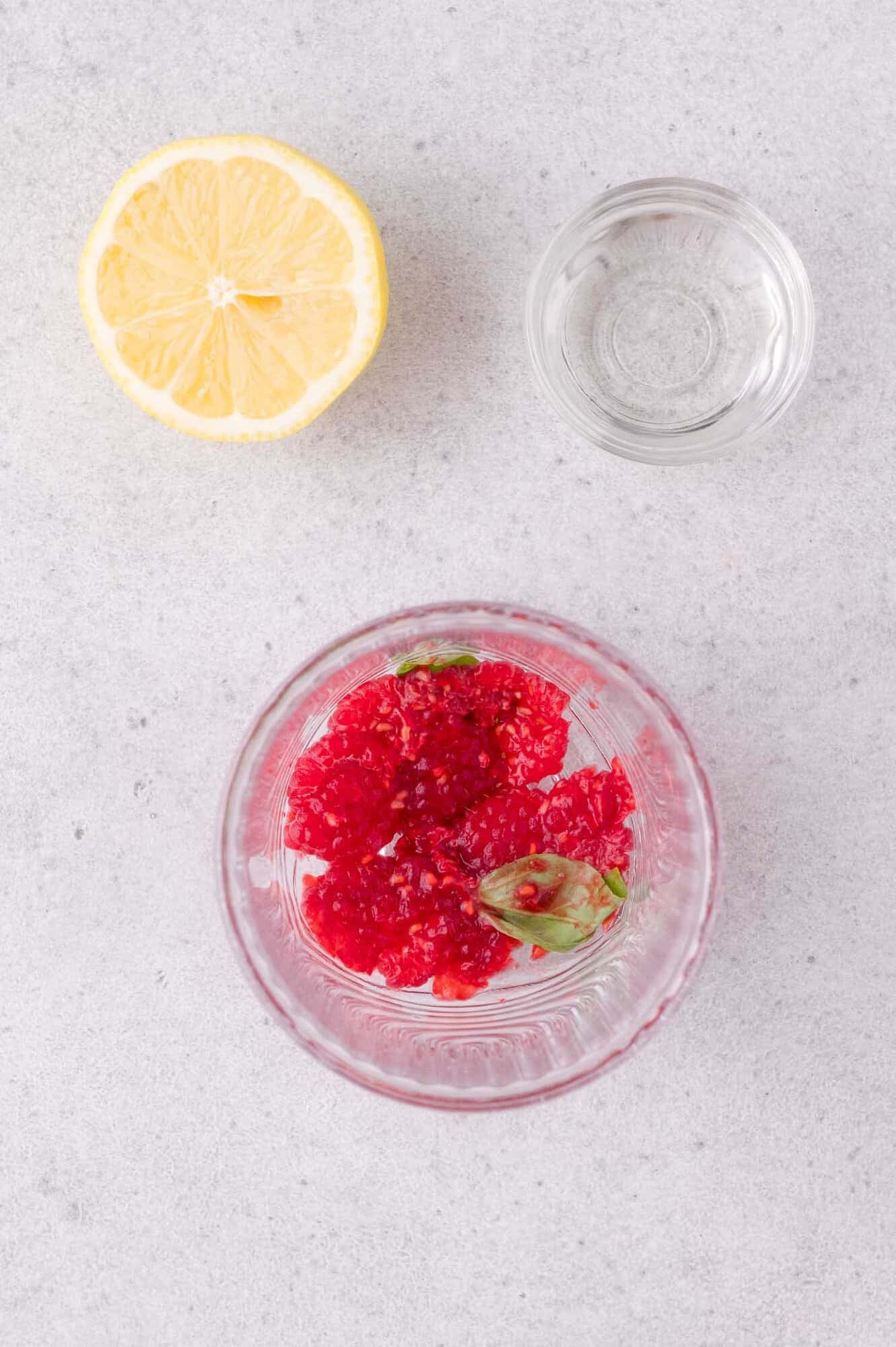 A glass with mashed raspberries and basil, a small bowl of clear liquid, and half a lemon on a light gray surface.
