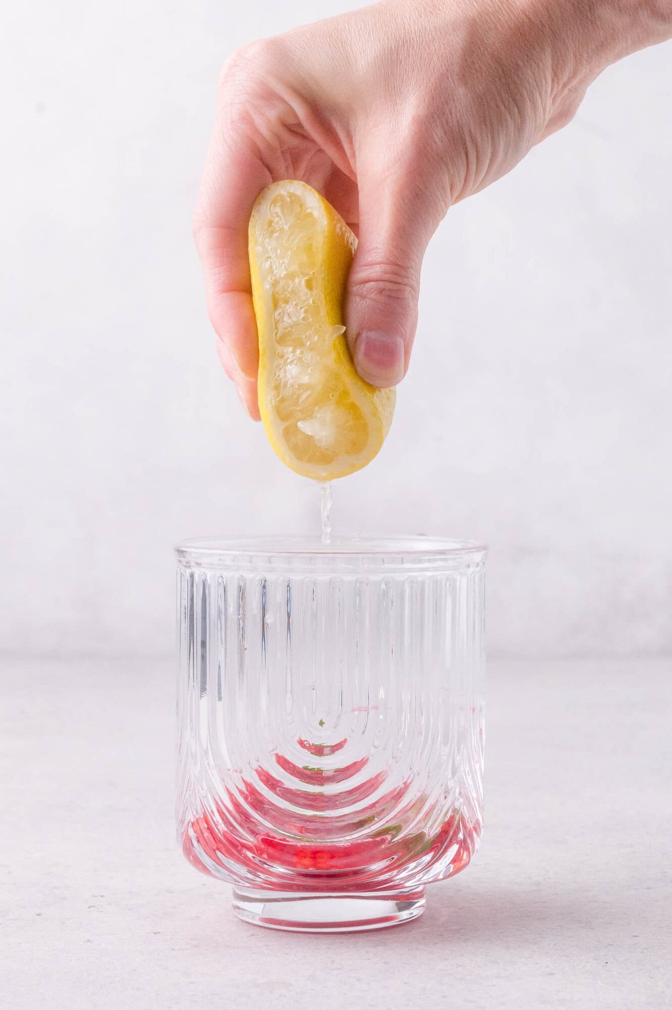 A hand squeezes half a lemon over a clear glass with a red liquid at the bottom, against a light background.