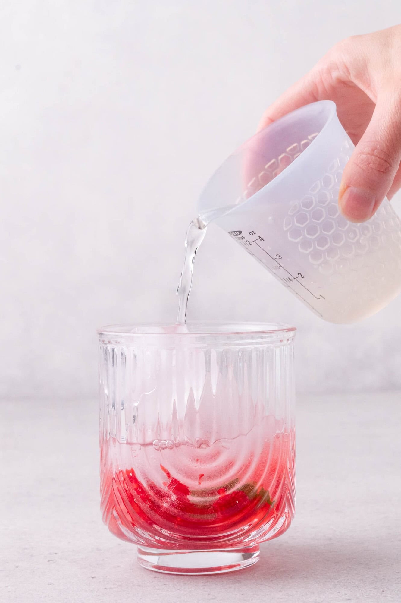 A hand pours sparkling water from a measuring cup into a glass containing red syrup and ice on a light surface.