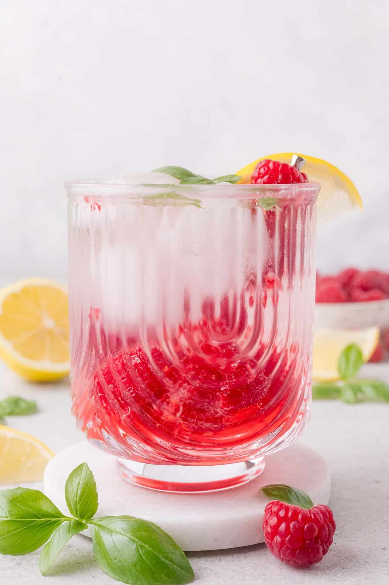 A glass of iced raspberry mocktail garnished with a lemon slice, fresh raspberries, and basil, sits on a white surface with lemon halves and basil leaves around it.