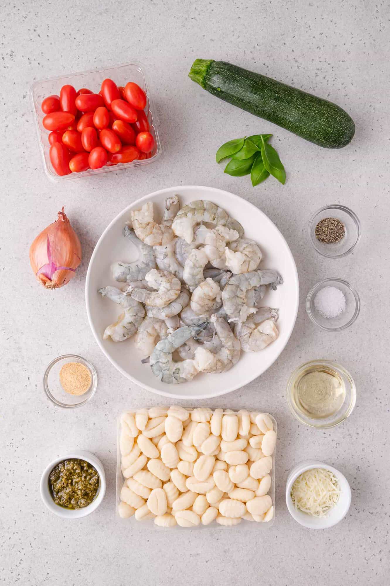 Ingredients for a shrimp and gnocchi dish arranged on a countertop: raw shrimp, gnocchi, cherry tomatoes, zucchini, shallot, basil, pesto, grated cheese, seasoning, and oil.