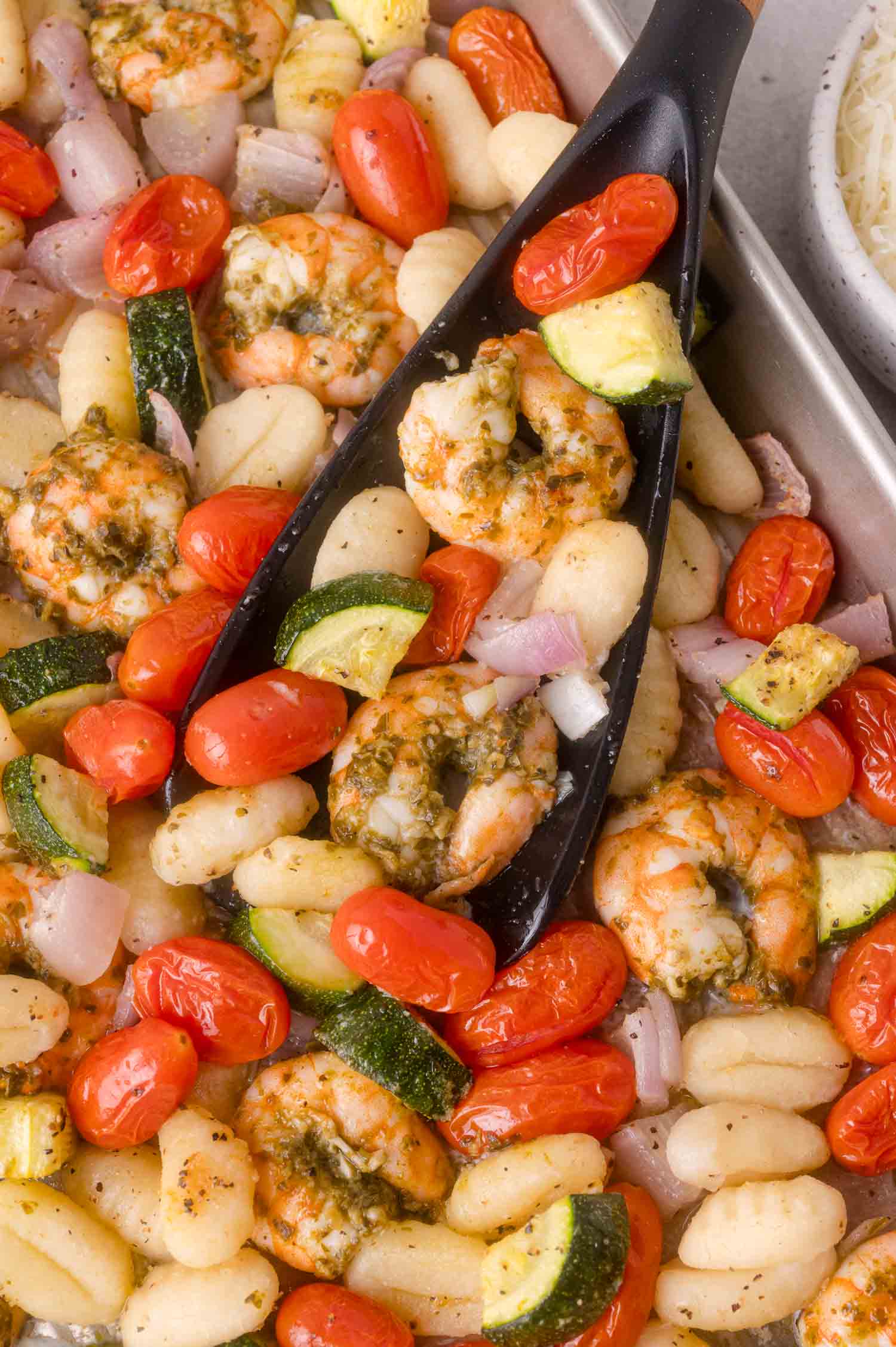 Sheet pan with cooked shrimp, gnocchi, cherry tomatoes, zucchini, and red onions, with a black serving spoon lifting a portion.