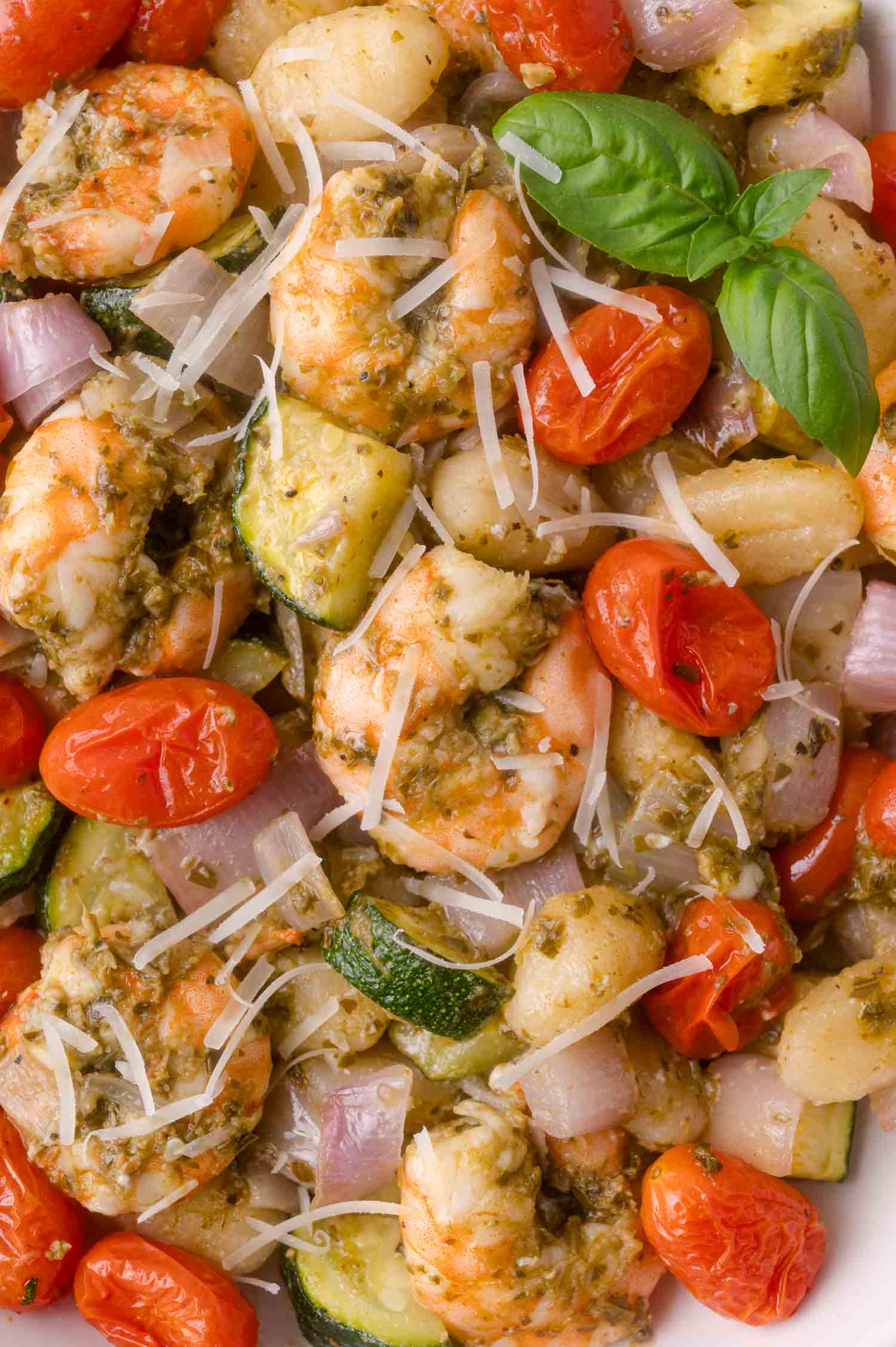 Cooked shrimp with cherry tomatoes, zucchini, onions, shredded cheese, and pesto, garnished with a basil sprig.