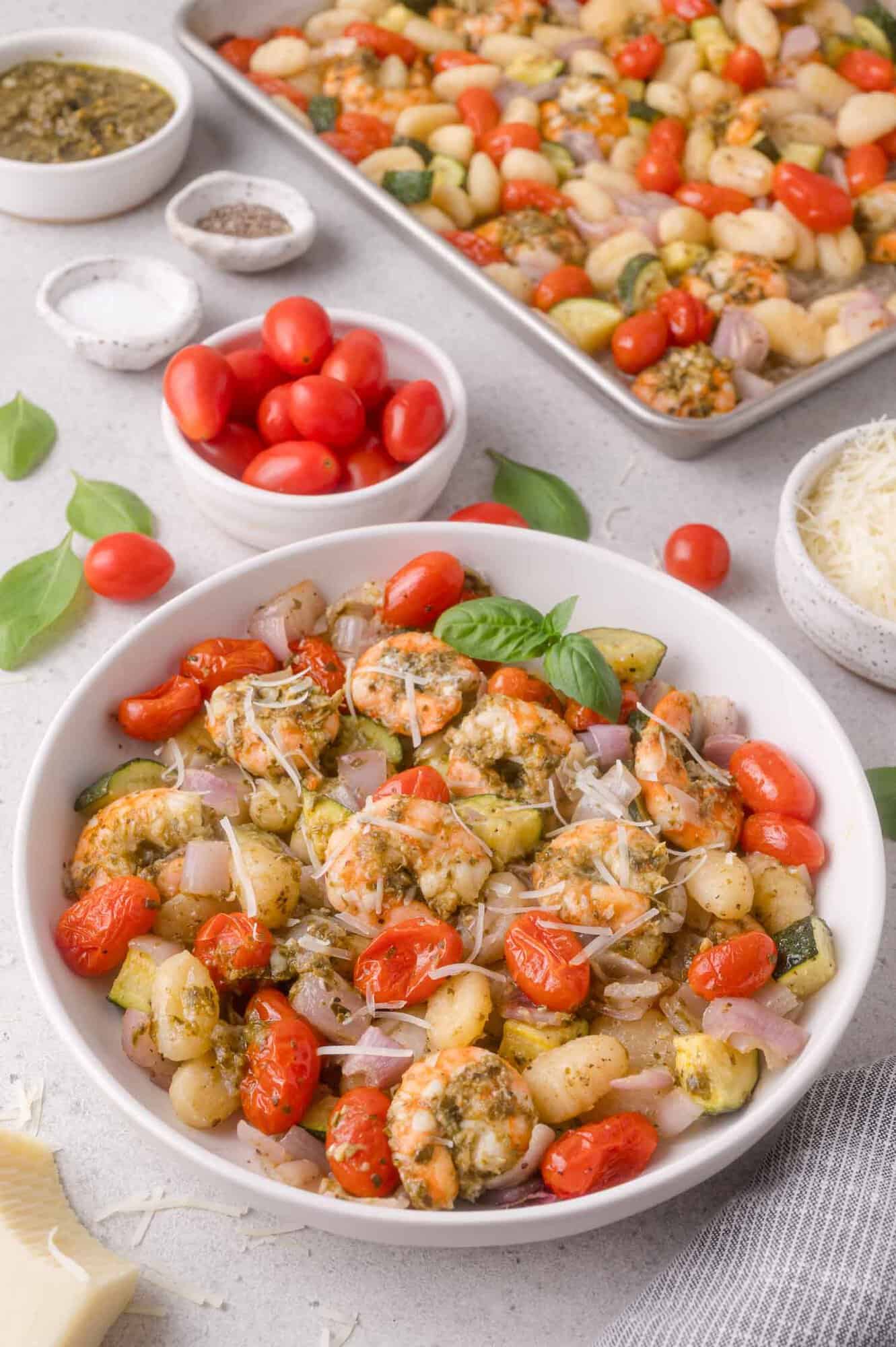 Bowl of gnocchi with shrimp, cherry tomatoes, zucchini, red onion, and herbs, with a tray of the same dish, grated cheese, and fresh tomatoes in the background.