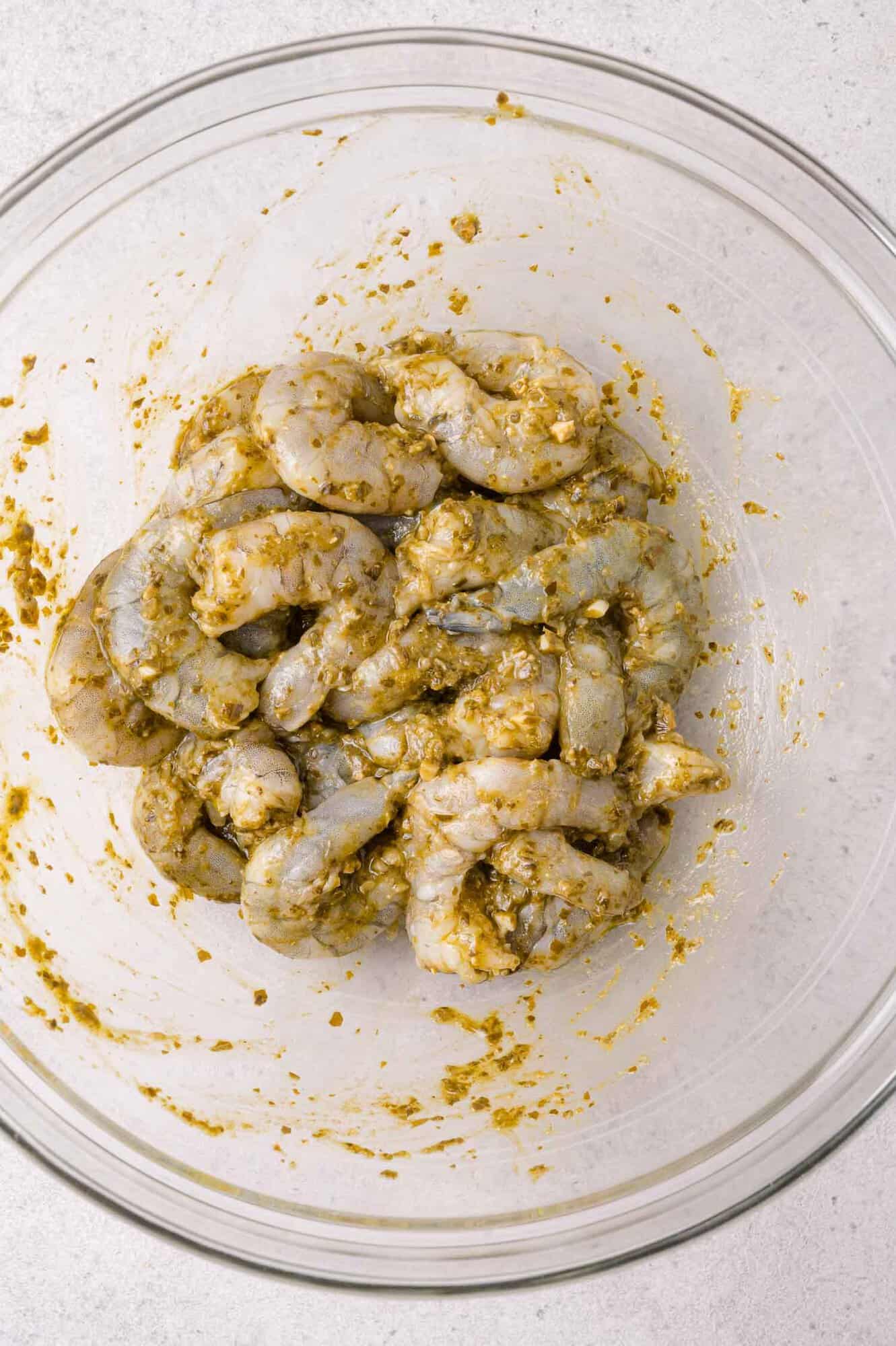 Raw shrimp marinating in pesto in a clear glass bowl on a light-colored surface.