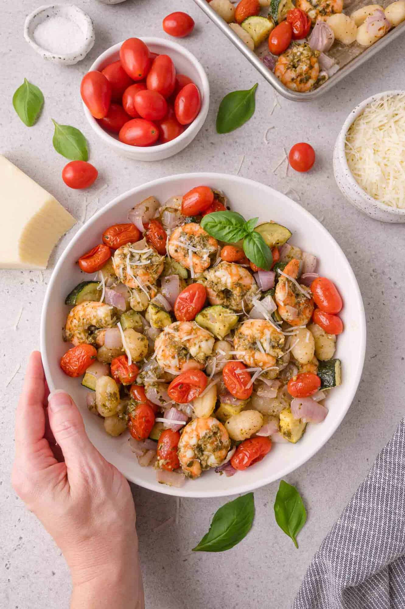A hand holds a bowl of baked gnocchi with tomatoes, zucchini, shrimp, and herbs. Nearby are parmesan cheese, grape tomatoes, basil leaves, and more gnocchi on a tray.