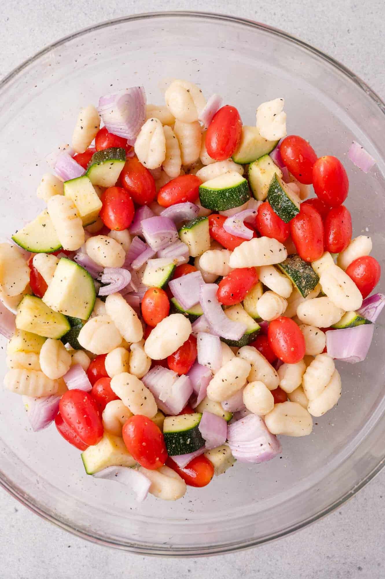 A glass bowl filled with uncooked gnocchi, halved cherry tomatoes, chopped zucchini, and diced red onions, seasoned with black pepper.