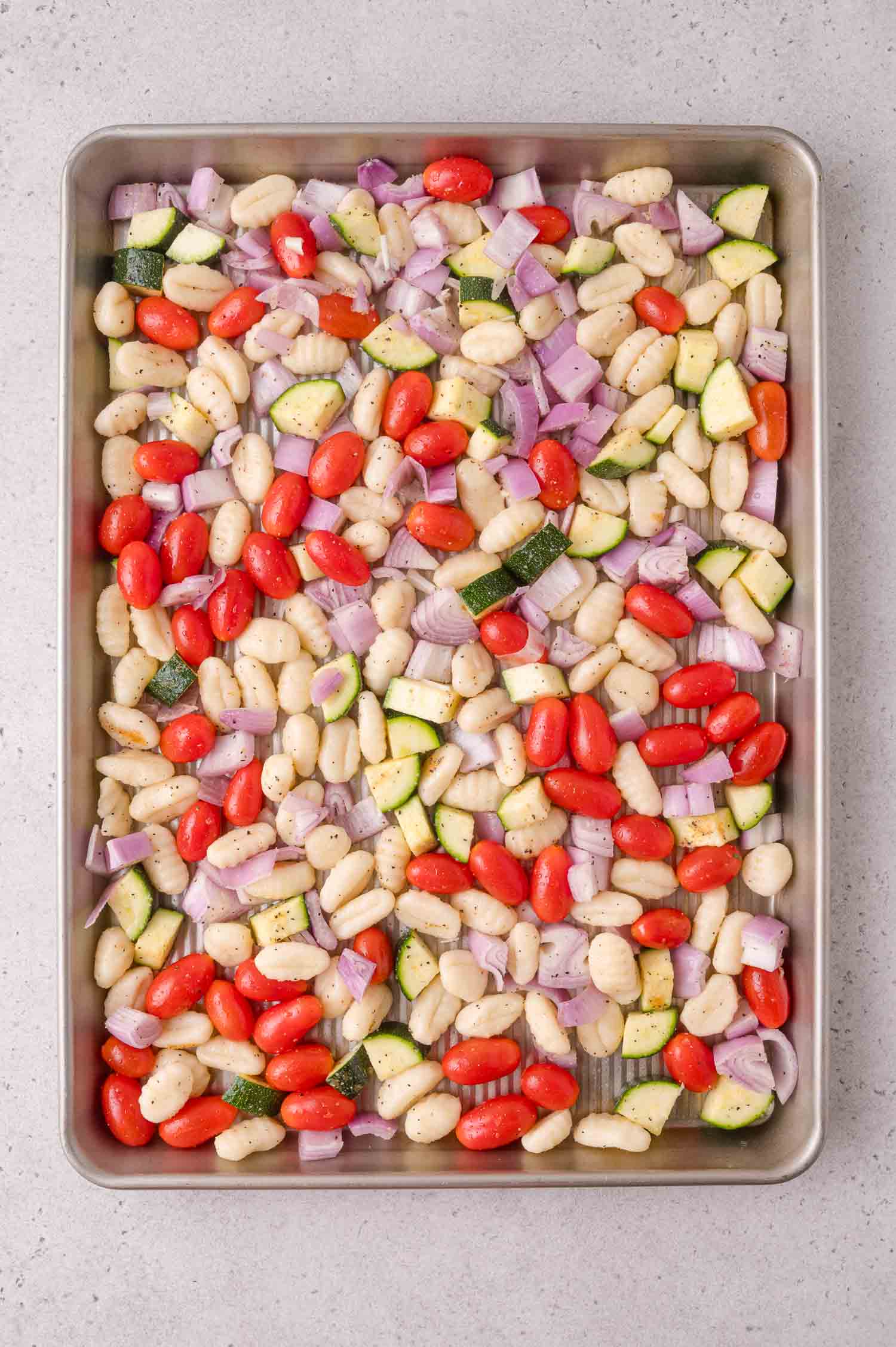 A baking sheet with uncooked gnocchi, cherry tomatoes, diced red onion, and zucchini, seasoned and ready to roast.