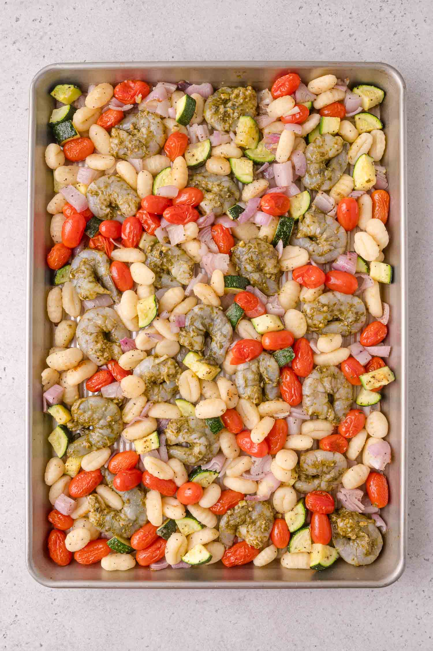Sheet pan with raw shrimp, gnocchi, cherry tomatoes, zucchini, red onion, and dollops of pesto, ready to be cooked.