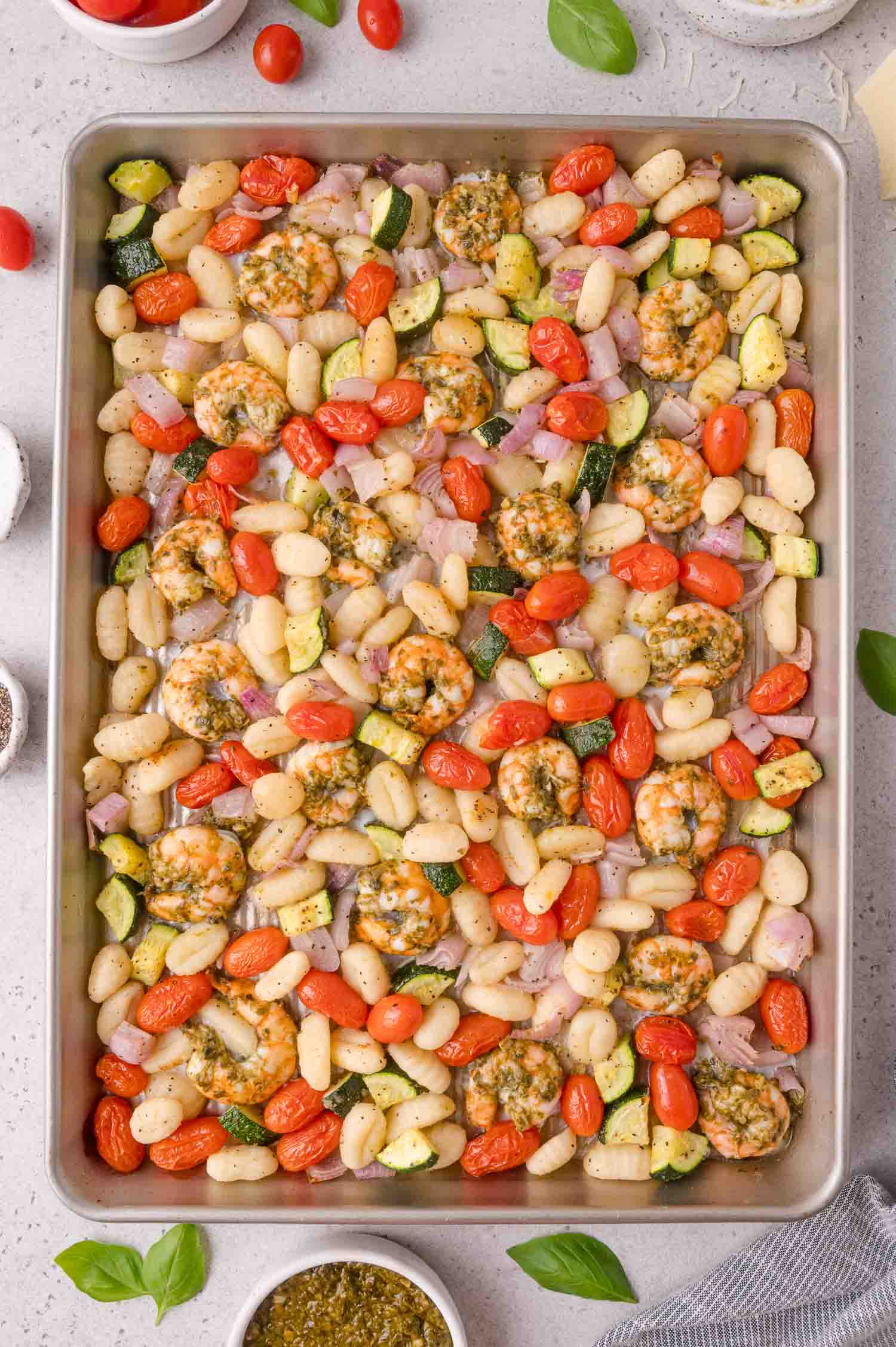 Sheet pan with cooked shrimp, gnocchi, cherry tomatoes, zucchini, and red onion, garnished with herbs. Small bowls of ingredients and basil leaves are visible around the tray.