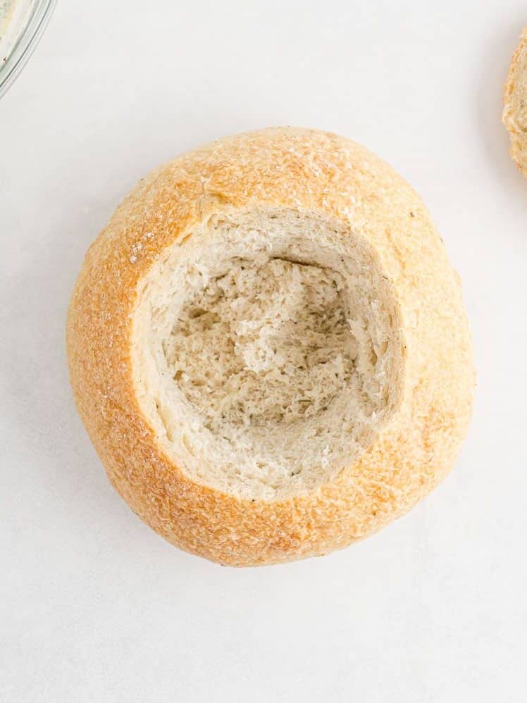 A round bread loaf with the top cut off and the inside hollowed out, placed on a white surface.