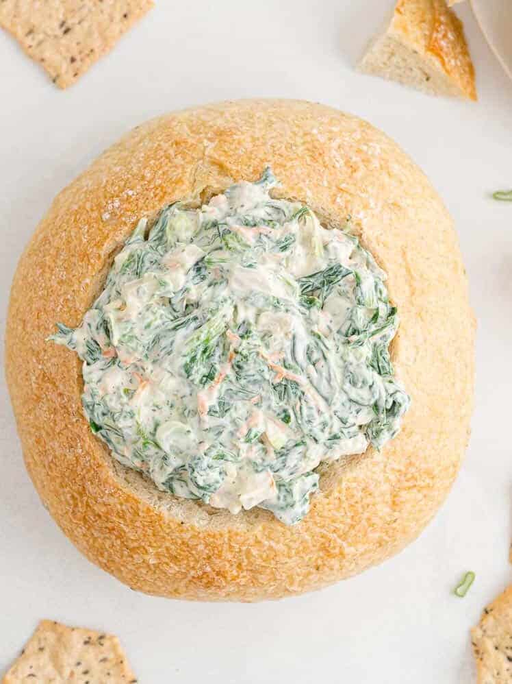 A round bread loaf filled with creamy spinach dip is shown from above, surrounded by pieces of crackers on a white surface.