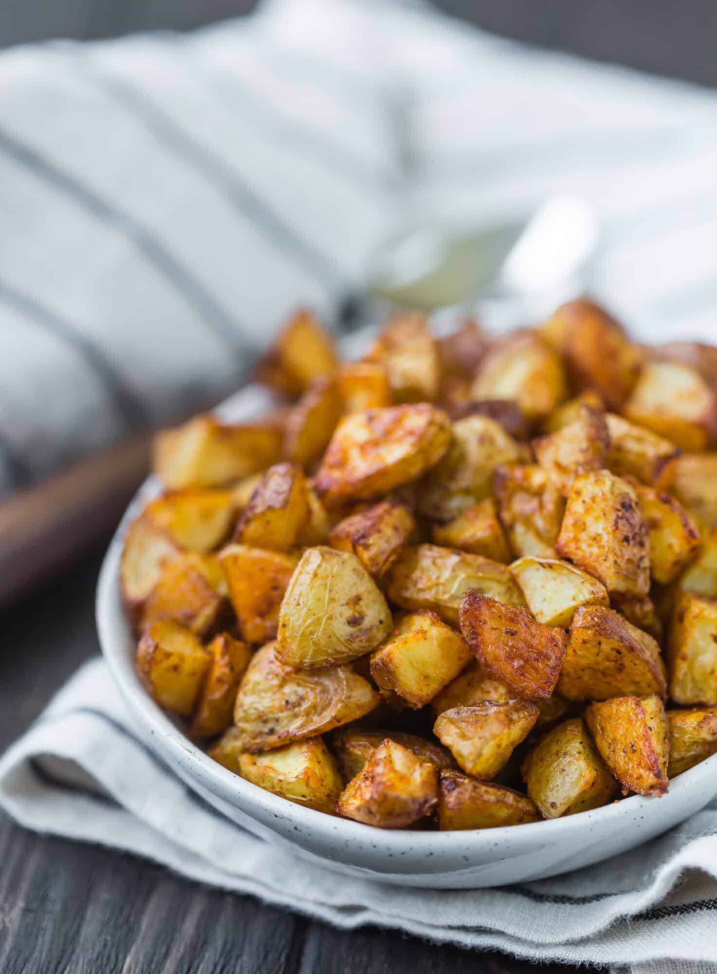 A bowl of roasted diced potatoes sits on a striped cloth with a blurred fork in the background.