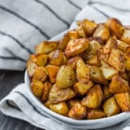 A white bowl filled with roasted, diced potatoes sits on a black and white striped cloth on a dark wooden surface.