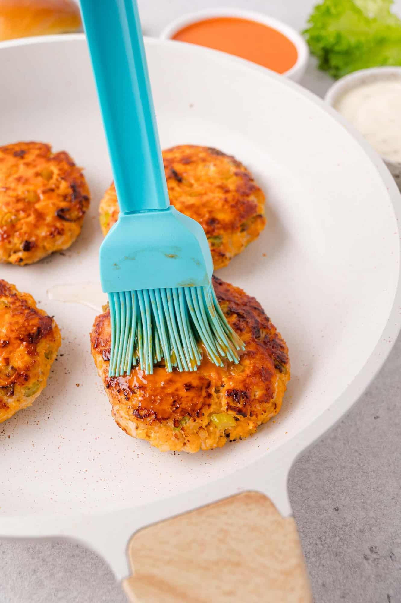 A blue silicone brush applies sauce to a cooked patty in a white skillet, with three other patties and bowls of sauces in the background.