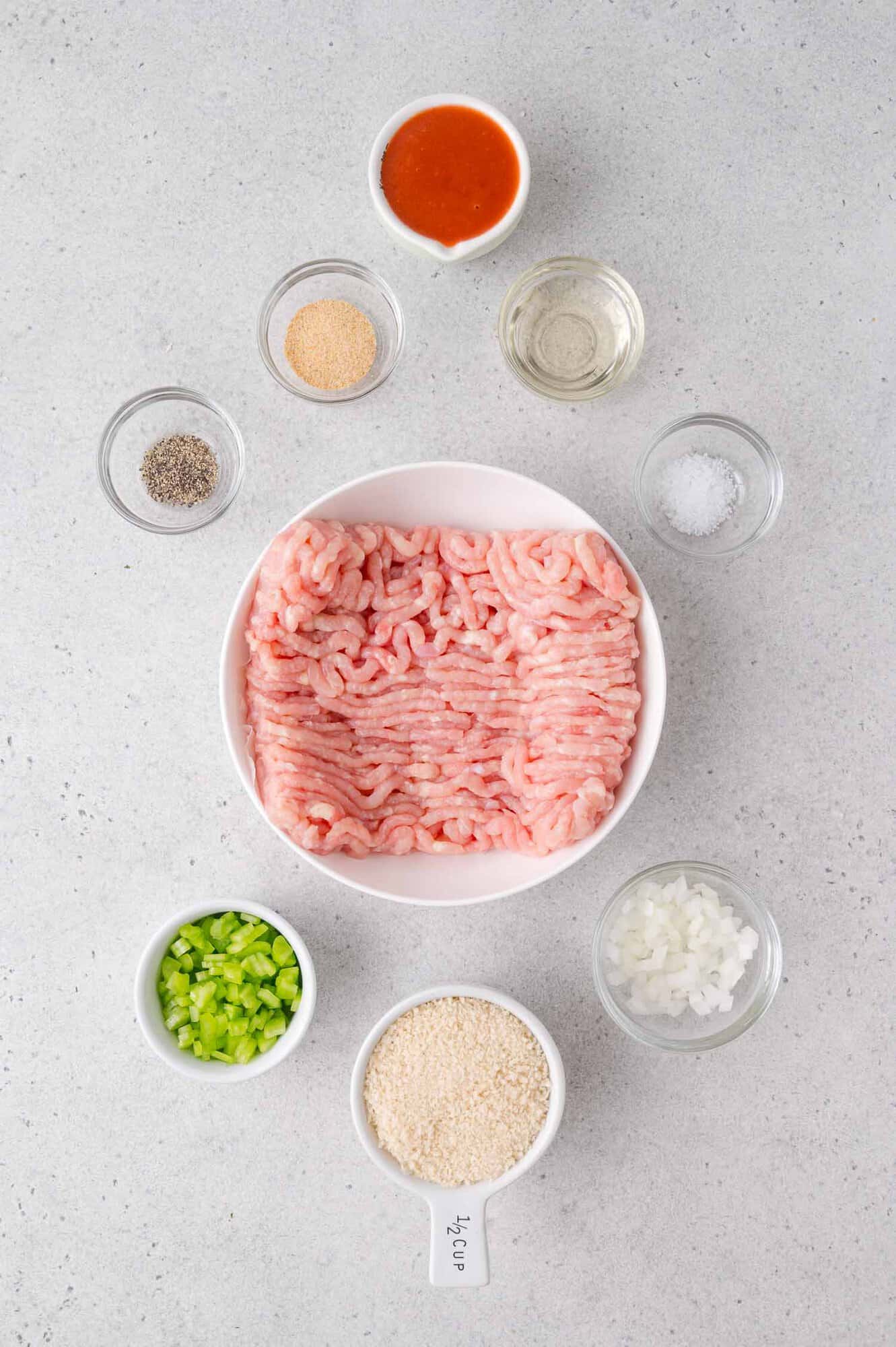 A plate of raw ground chicken is surrounded by small bowls of diced onion, diced celery, breadcrumbs, pepper, garlic powder, salt, hot sauce, and oil on a light countertop.