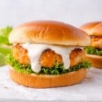A buffalo chicken burger with lettuce and white sauce is served on a round bun, placed on white parchment paper, with a second burger in the background.