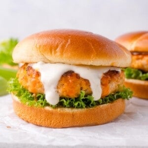 A buffalo chicken burger with lettuce and white sauce is served on a round bun, placed on white parchment paper, with a second burger in the background.