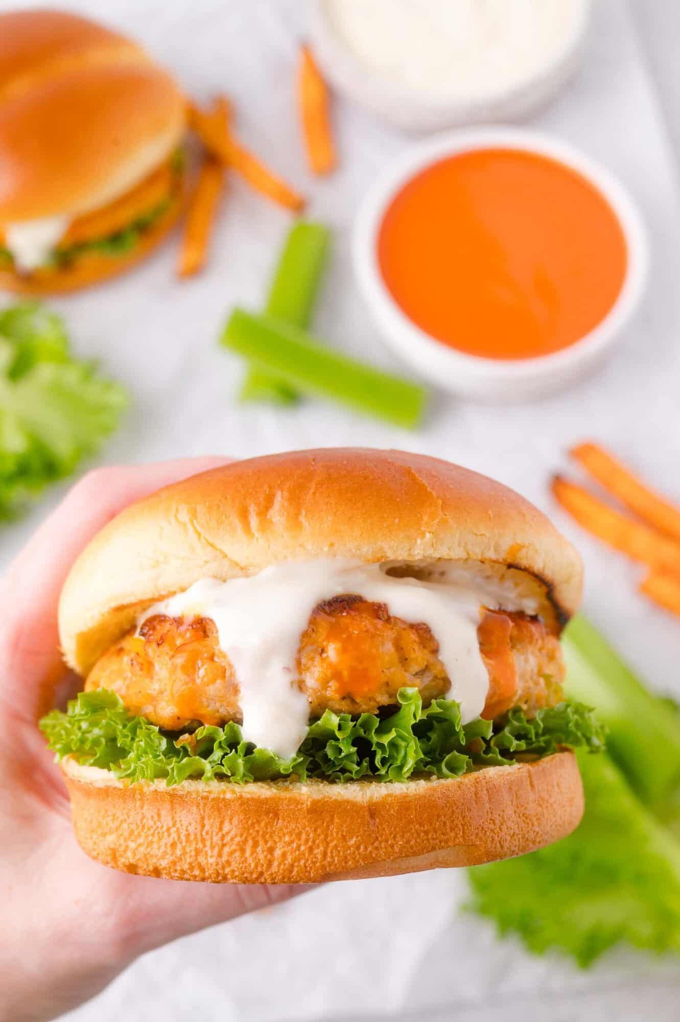 A hand holds a chicken burger with lettuce and ranch sauce, with celery sticks, and dipping sauces in the background.
