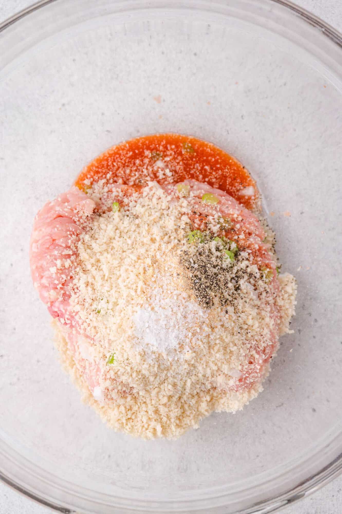 Ground meat, breadcrumbs, seasoning, and sauce in a glass mixing bowl, ready to be combined for a recipe.