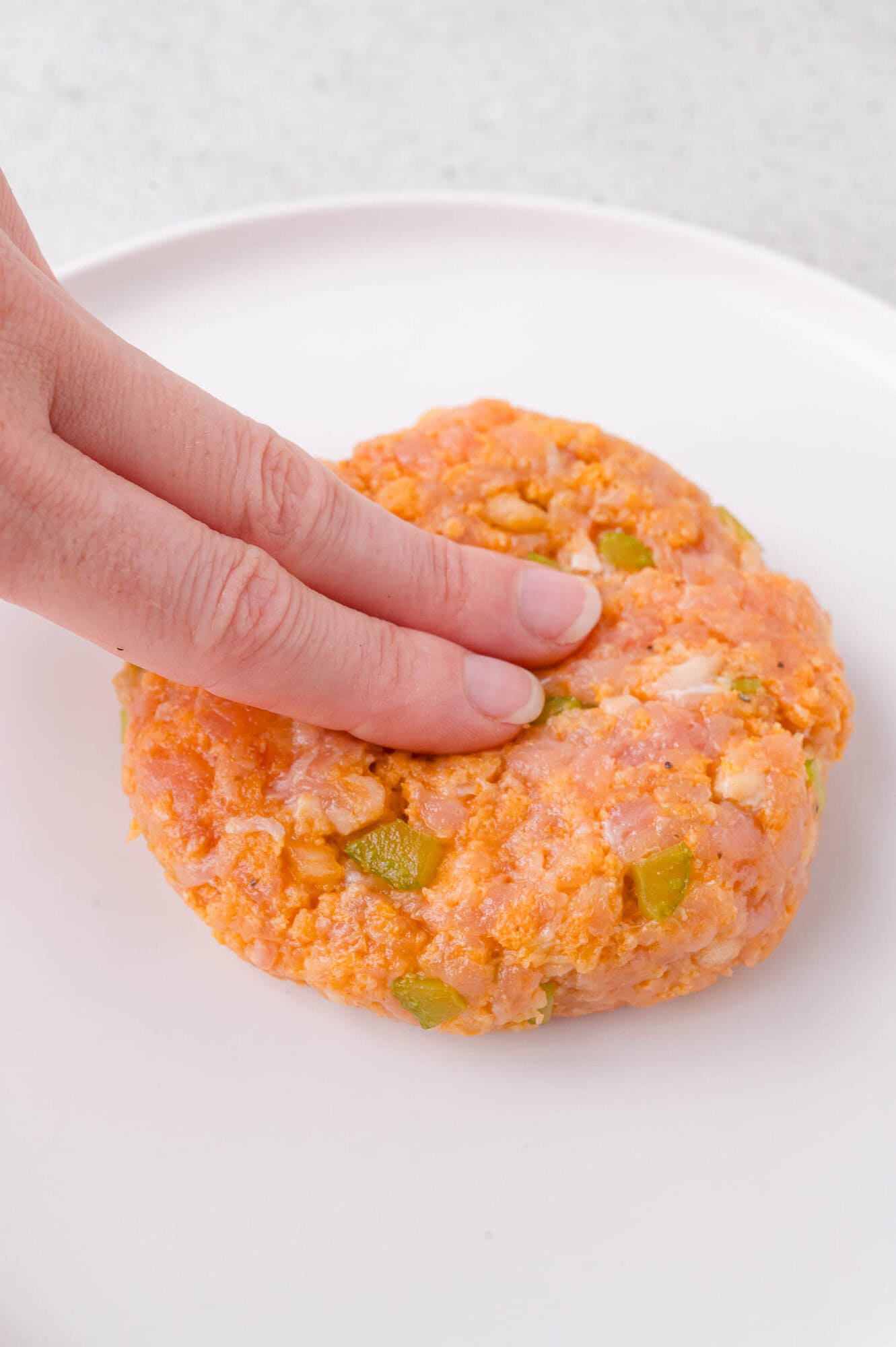 A hand presses down on a raw chicken burger mixed with chopped vegetables on a white plate.