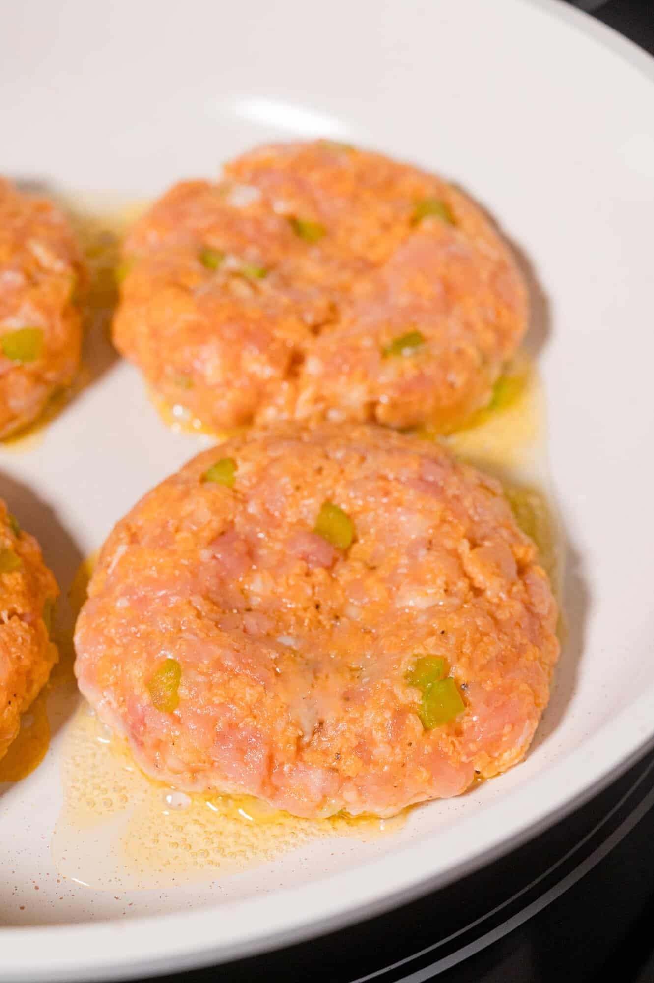 Raw seasoned ground chicken patties with visible celery pieces are arranged in a white skillet with some oil.