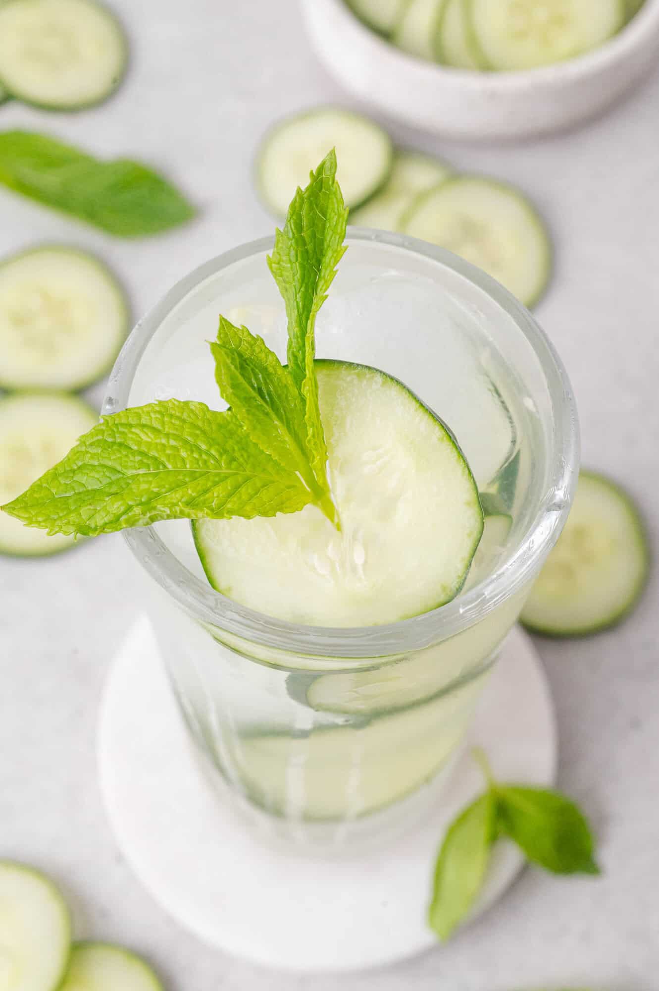 A glass of water with ice, garnished with a cucumber slice and a fresh mint sprig, sits on a white coaster with cucumber slices and mint leaves scattered around.