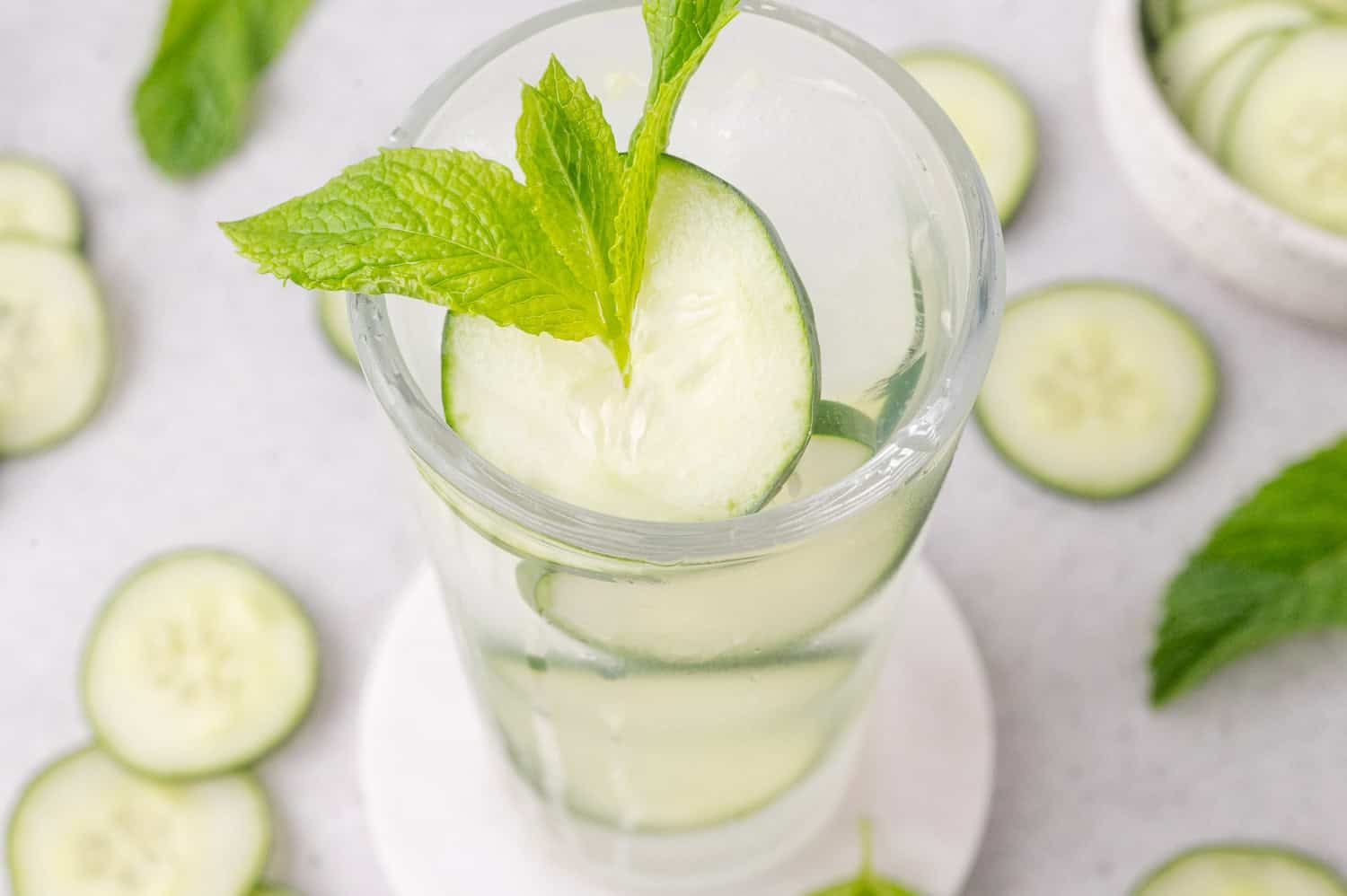 A glass of cucumber lime cooler with cucumber slices and a sprig of mint, placed on a white coaster, surrounded by cucumber slices and mint leaves.