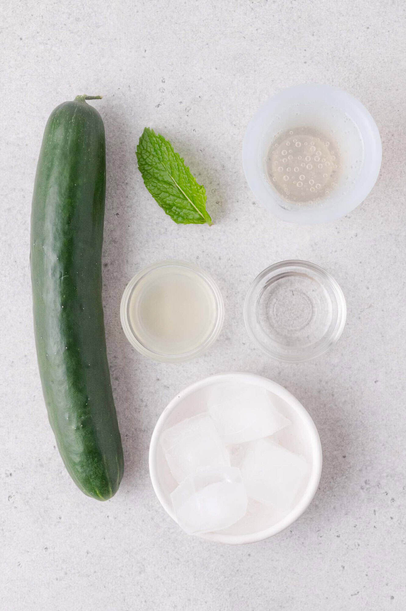 Cucumber, mint leaf, ice cubes in a bowl, and three small bowls containing clear liquids arranged on a light gray surface.