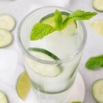 A glass of iced water garnished with cucumber slices and a sprig of mint, placed on a coaster with cucumber and lime slices in the background.
