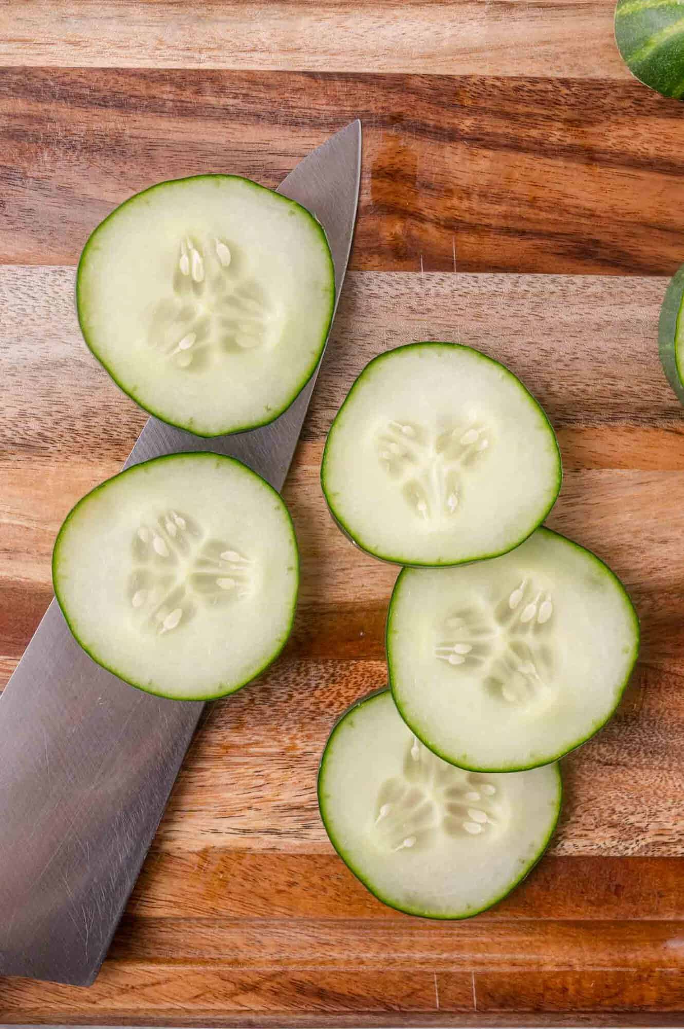 Five cucumber slices and a kitchen knife are on a wooden cutting board.