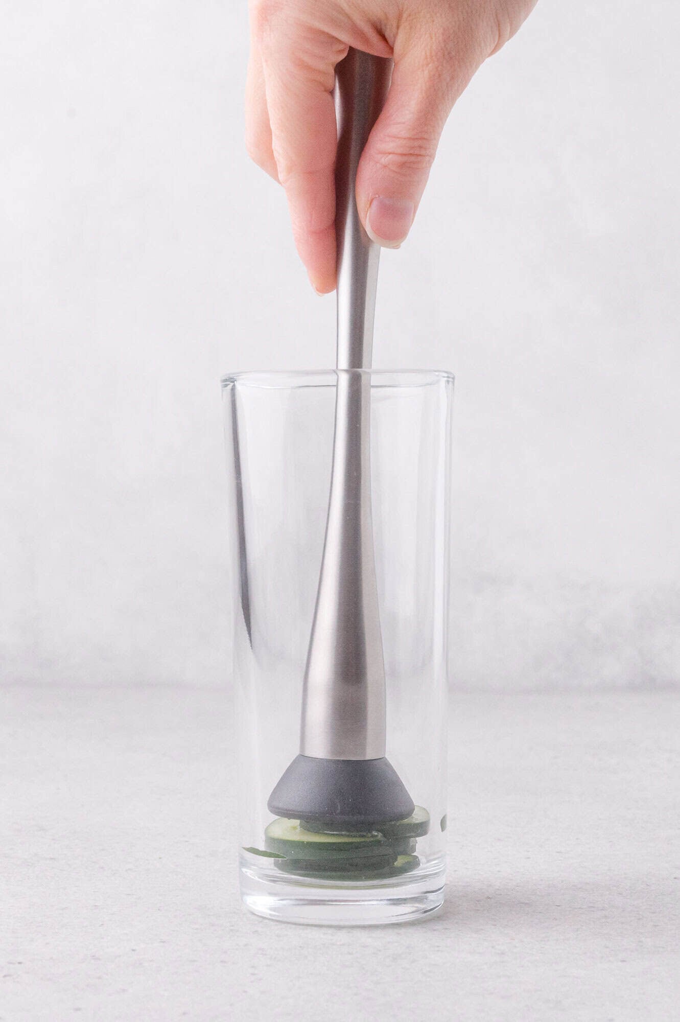 A hand uses a metal muddler to crush lime slices at the bottom of a clear glass on a light surface.
