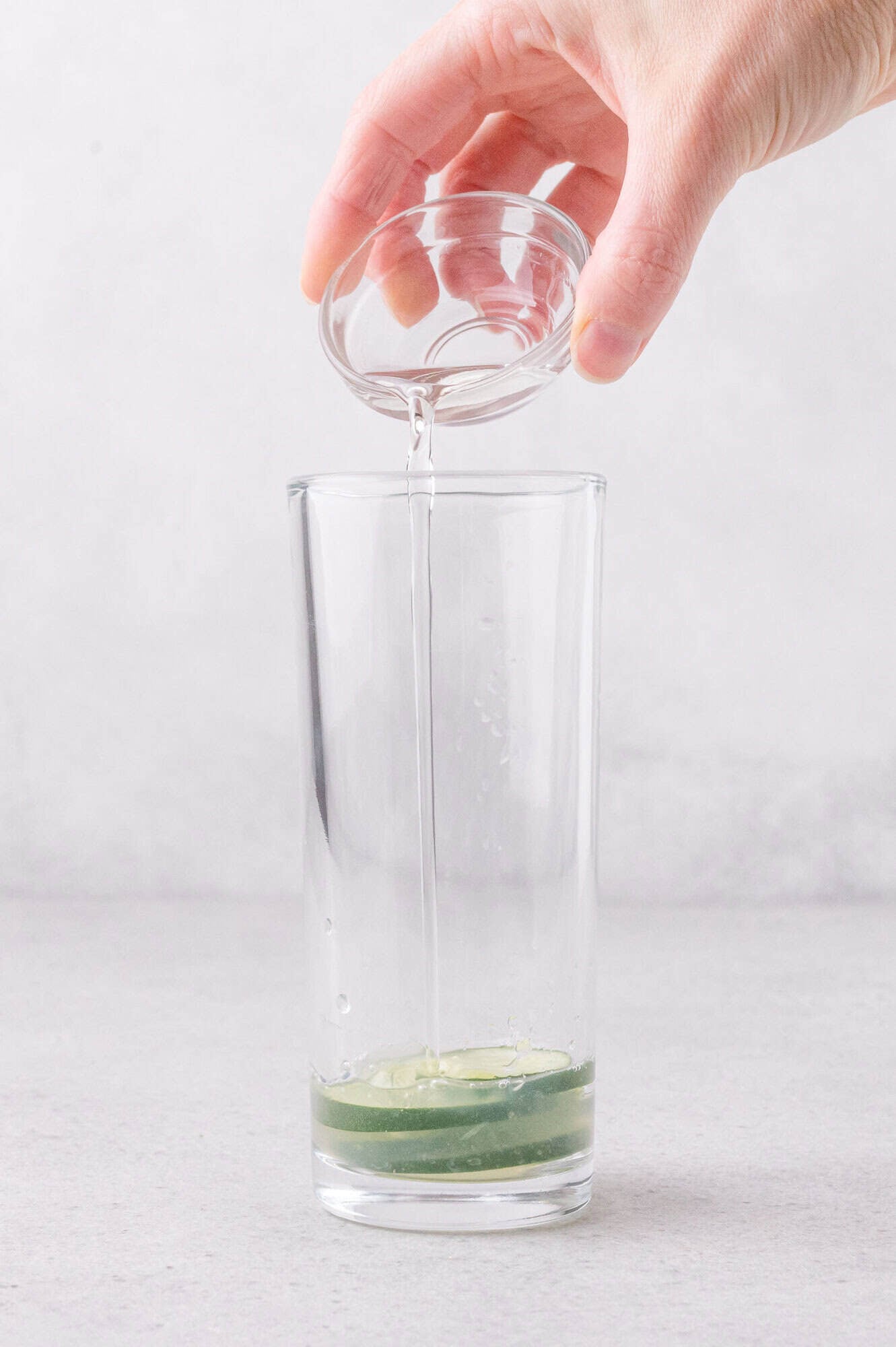A hand pours clear liquid from a small glass bowl into a tall glass containing cucumber slices and juice.