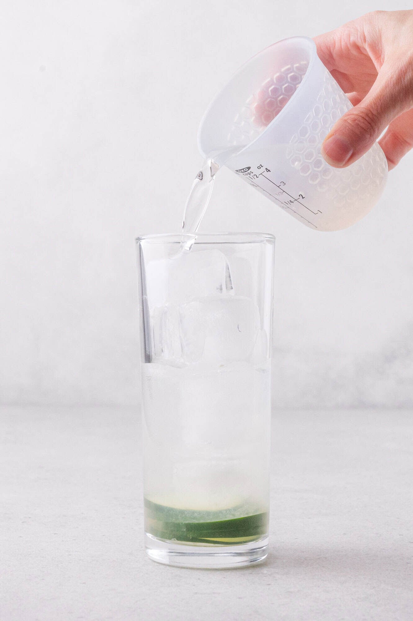 A hand pours clear liquid from a measuring cup into a tall glass filled with ice and a small amount of cucumbers at the bottom.