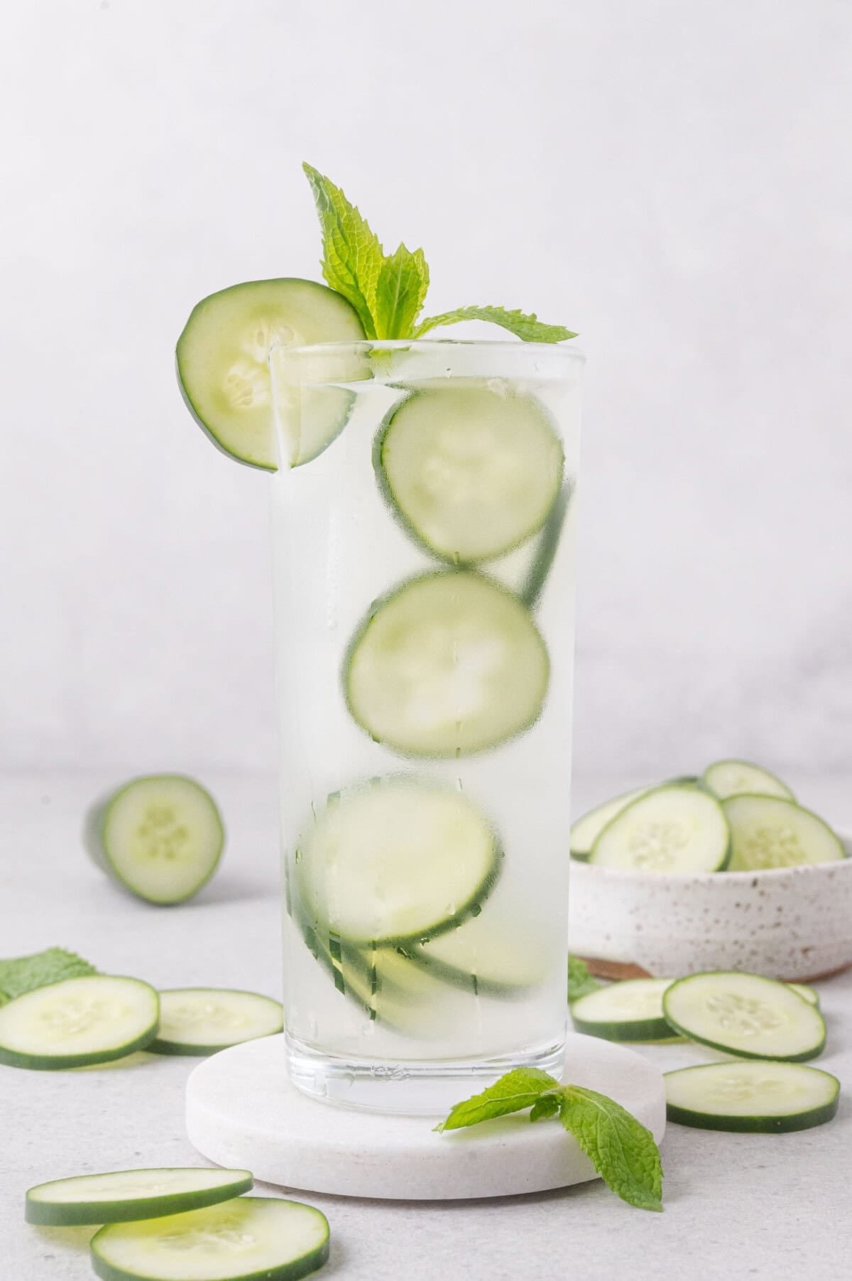 A tall glass of cucumber lime cooler with ice, cucumber slices, and a sprig of mint, surrounded by cucumber slices on a light surface.
